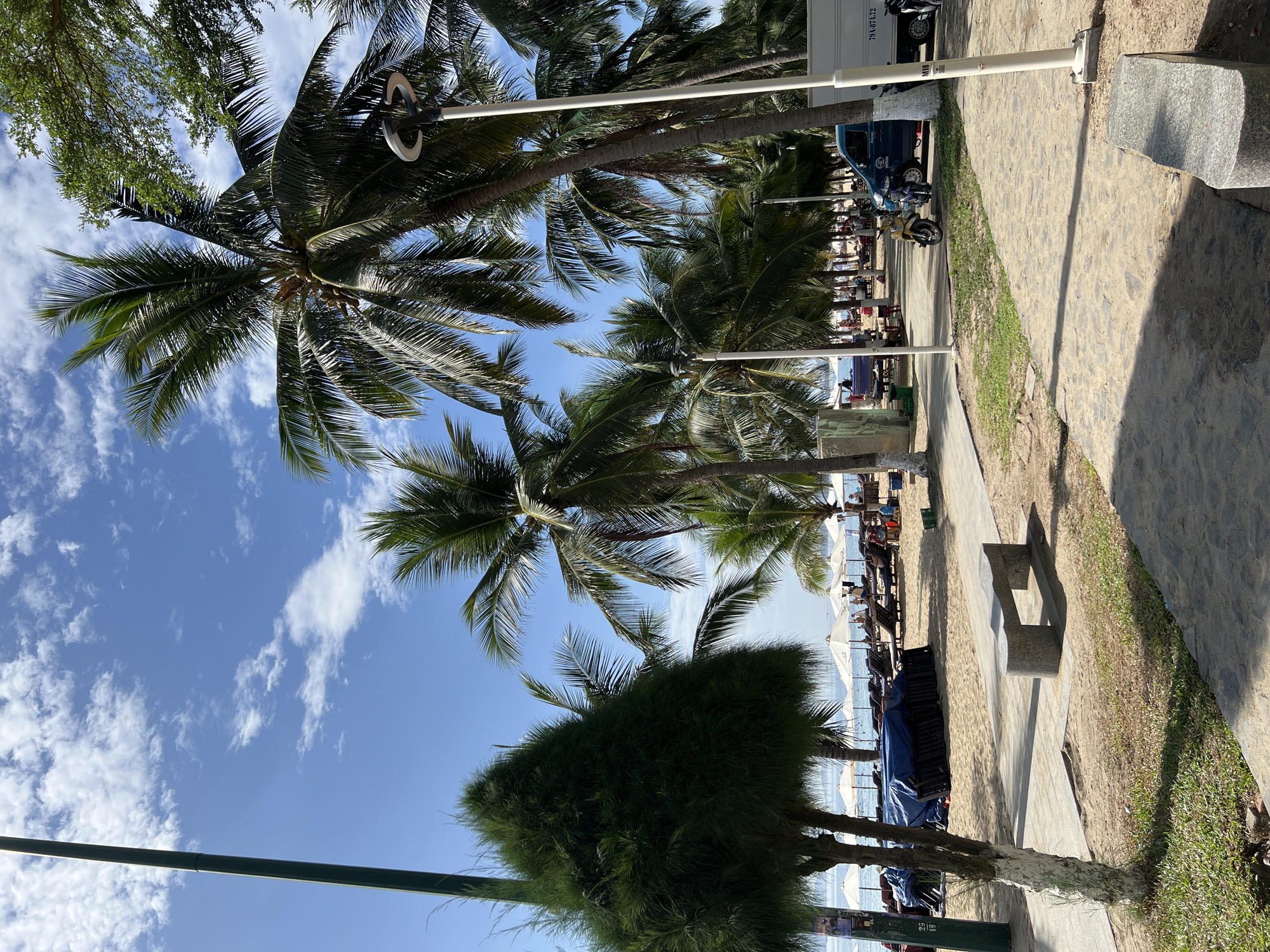 The image depicts a view of a tropical beach in Nha Trang, Vietnam. Tall palm trees line the sandy shore, with their fronds reaching towards a bright blue sky dotted with white clouds. In the foreground, a paved walkway runs alongside the beach, bordered by patches of grass and sand. A gray stone bench is situated on the walkway, offering a place to rest.
Further back, beach chairs and umbrellas are arranged on the sand, with some people lounging and others appearing to be walking or interacting. A blue van and a yellow scooter are parked on the side of the walkway, near a white truck. The ocean is visible in the distance, with gentle waves lapping at the shore. The overall atmosphere suggests a sunny day at a popular tourist destination.