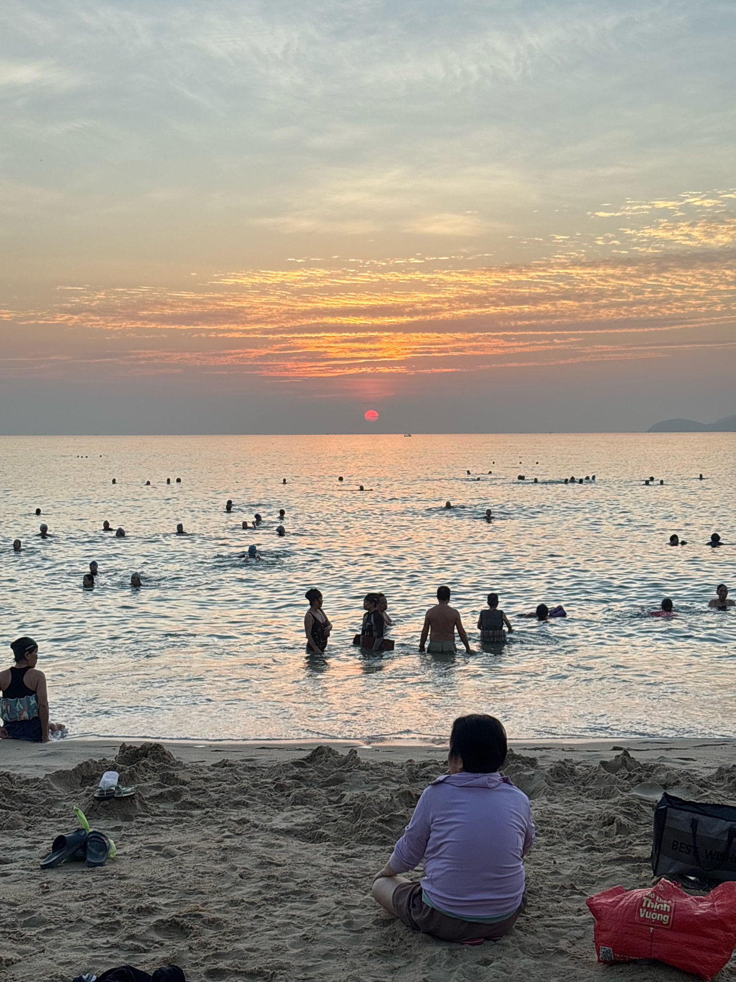 The image depicts a sunset scene at a beach in Nha Trang, Vietnam. The sky is filled with orange and pink hues, with wispy clouds. The sun is a small, red orb low on the horizon, casting a warm glow on the water. Numerous people are swimming and wading in the shallow ocean, with some further out in the water. On the sandy beach in the foreground, a person is sitting facing away from the camera, wearing a purple top and brown shorts. Nearby are several bags, a pair of flip-flops, and snorkel gear. The overall atmosphere is tranquil and suggests a pleasant evening at the beach.
