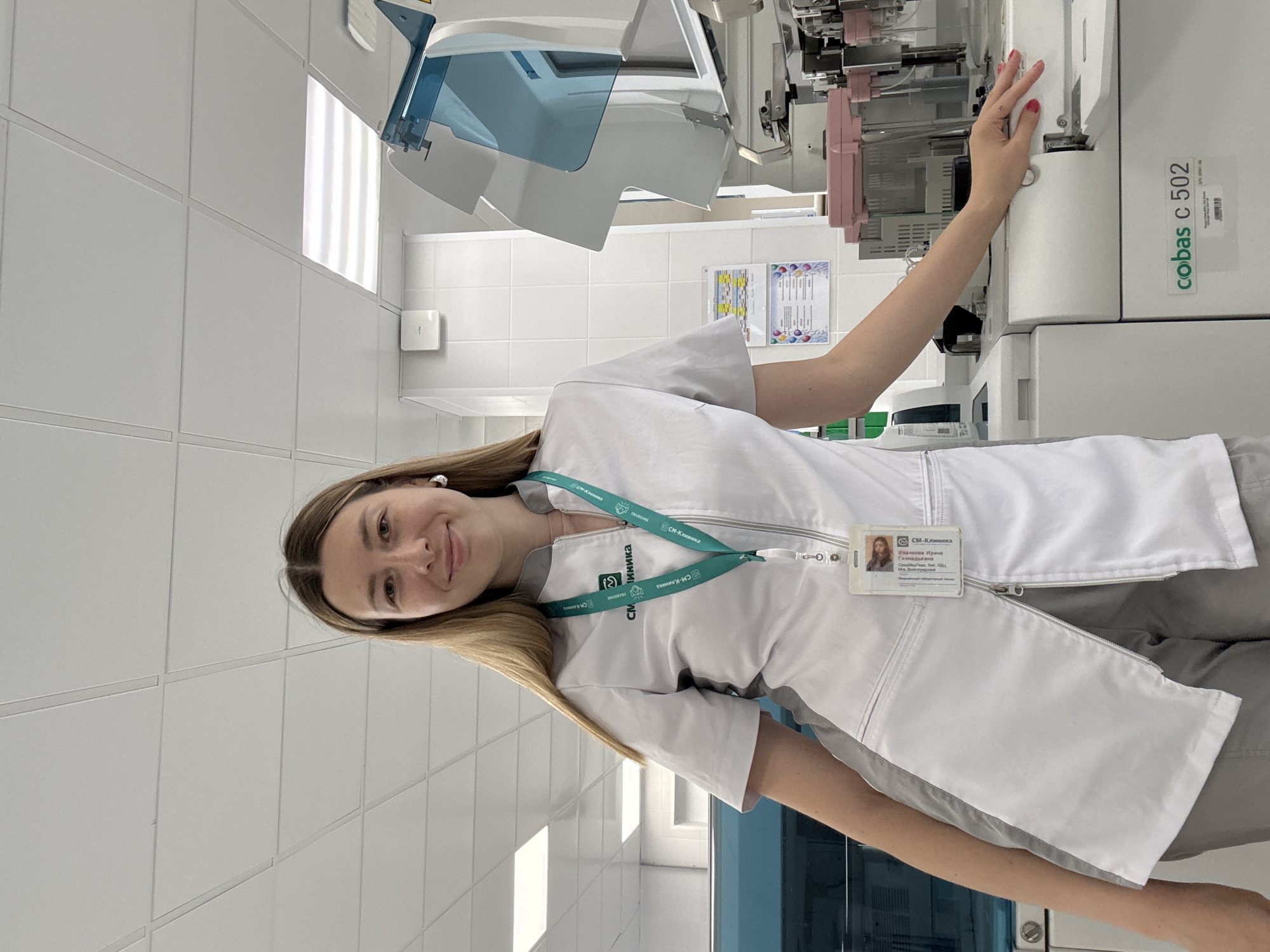 A medium shot captures a fair-skinned woman with long, straight blonde hair, wearing a light gray zip-up lab coat with a green lanyard featuring a logo. She is smiling and looking at the camera. The background appears to be a laboratory setting, with white tiled walls and ceiling panels. Visible equipment includes a large white machine labeled "cobas c 502" and other laboratory instruments. A name tag with a photo is attached to her coat. The lighting is bright and even, suggesting an indoor environment. The image does not provide any specific visual cues that would indicate a location within the city of Moscow, Russia.