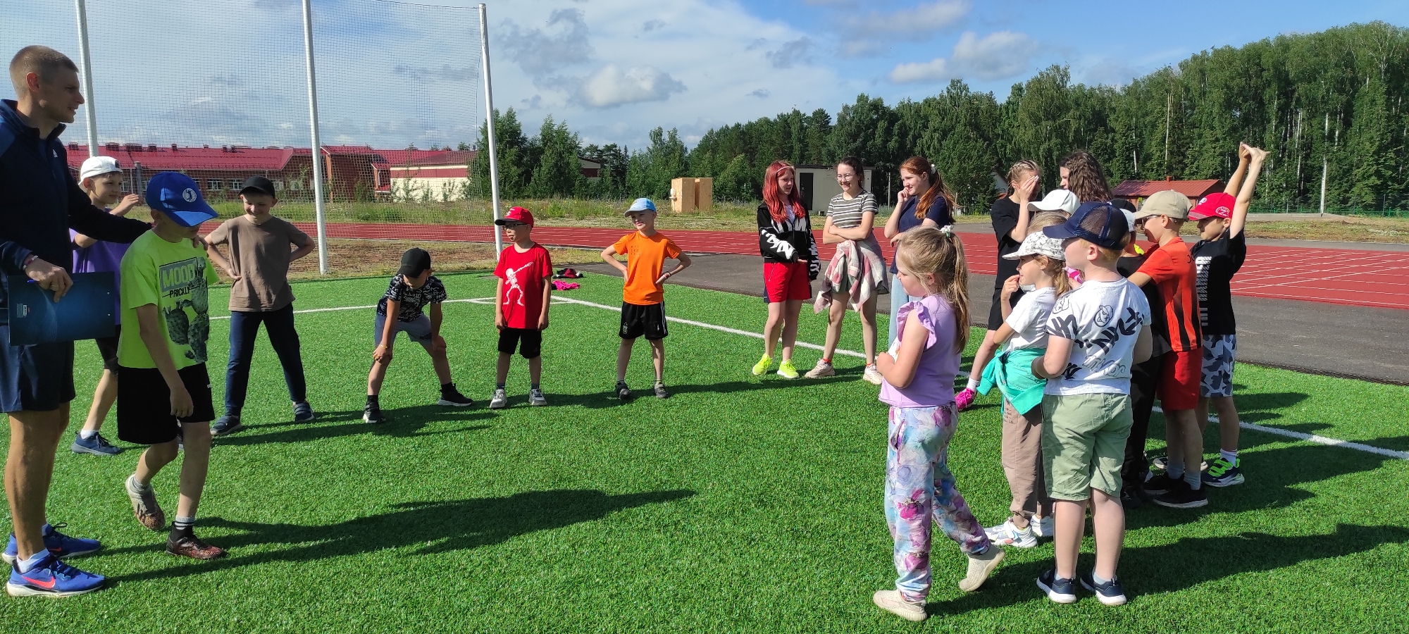 An outdoor group of children and two adults are gathered on a bright green artificial turf field, likely a sports ground, under a partly cloudy sky. The field is marked with white lines. In the background, a red running track and a treeline are visible, suggesting the location is near a wooded area or park. To the left, a red building with a white roof stands, and a tall fence or net structure is present.
On the left, a male adult, likely a coach, wearing a dark blue jacket over a t-shirt and dark shorts, holds a clipboard. Beside him, several children stand in a loose group. One boy wears a lime green t-shirt with a graphic print and black shorts, with a blue baseball cap. Another boy, to his right, wears a brown t-shirt and dark trousers. A child in a red t-shirt and black shorts, with a red cap, stands further to the right. An orange t-shirt is worn by a boy with black shorts and a light blue cap.
In the center and to the right of the image, a larger group of children are interacting. Some children have red hair, and several are wearing casual clothing such as t-shirts, shorts, and patterned trousers. One young girl in the foreground wears a purple t-shirt and patterned trousers. Another child is seen with their hands raised. The children appear to be engaged in some form of organized activity or game.
The overall atmosphere is bright and appears to be during daylight hours. The image captures a moment of activity and social interaction among a group of children and adults outdoors.
The location is identified as Noviy, Russia, likely on a sports facility or park within the area.
