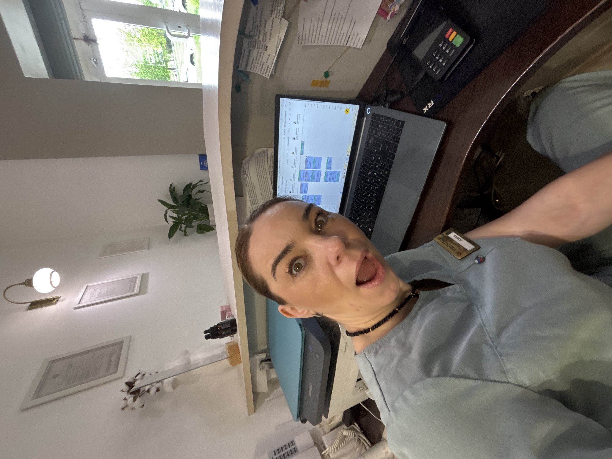 The image is taken from the perspective of a person in what appears to be a medical office setting. The person, likely a staff member based on their attire (a light blue uniform with a name tag that reads "Anna"), is making a surprised facial expression with their mouth open.
In the background, there's a reception desk with a laptop displaying a calendar or scheduling application. There's also a payment terminal on the desk, along with some paperwork.
The office environment is suggested by the framed documents on the wall, a small plant, and what seems to be a printer/scanner. The location is Unknown, Unknown.
