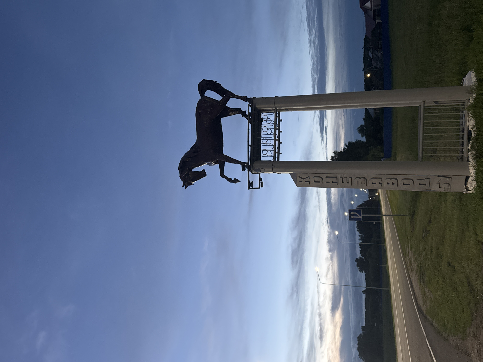 The image depicts a twilight scene in Nikolskoye, Russia, featuring a monument of a horse on a pedestal. The monument is positioned near a road with streetlights lining its side. The sky is a deep blue with scattered clouds, indicative of dusk. The horse sculpture is dark in color and poised with one leg raised. The pedestal is a light gray structure with vertical text that appears to read "КОНЕЗАВОДСКИЙ" which translates to "Stud Farm". The road is paved and curves to the left, bordered by green grass on both sides. In the background, trees and residential buildings are visible. There are no people or animals in the immediate vicinity of the monument or on the road. The overall atmosphere is calm and serene, characteristic of the transition from day to night.