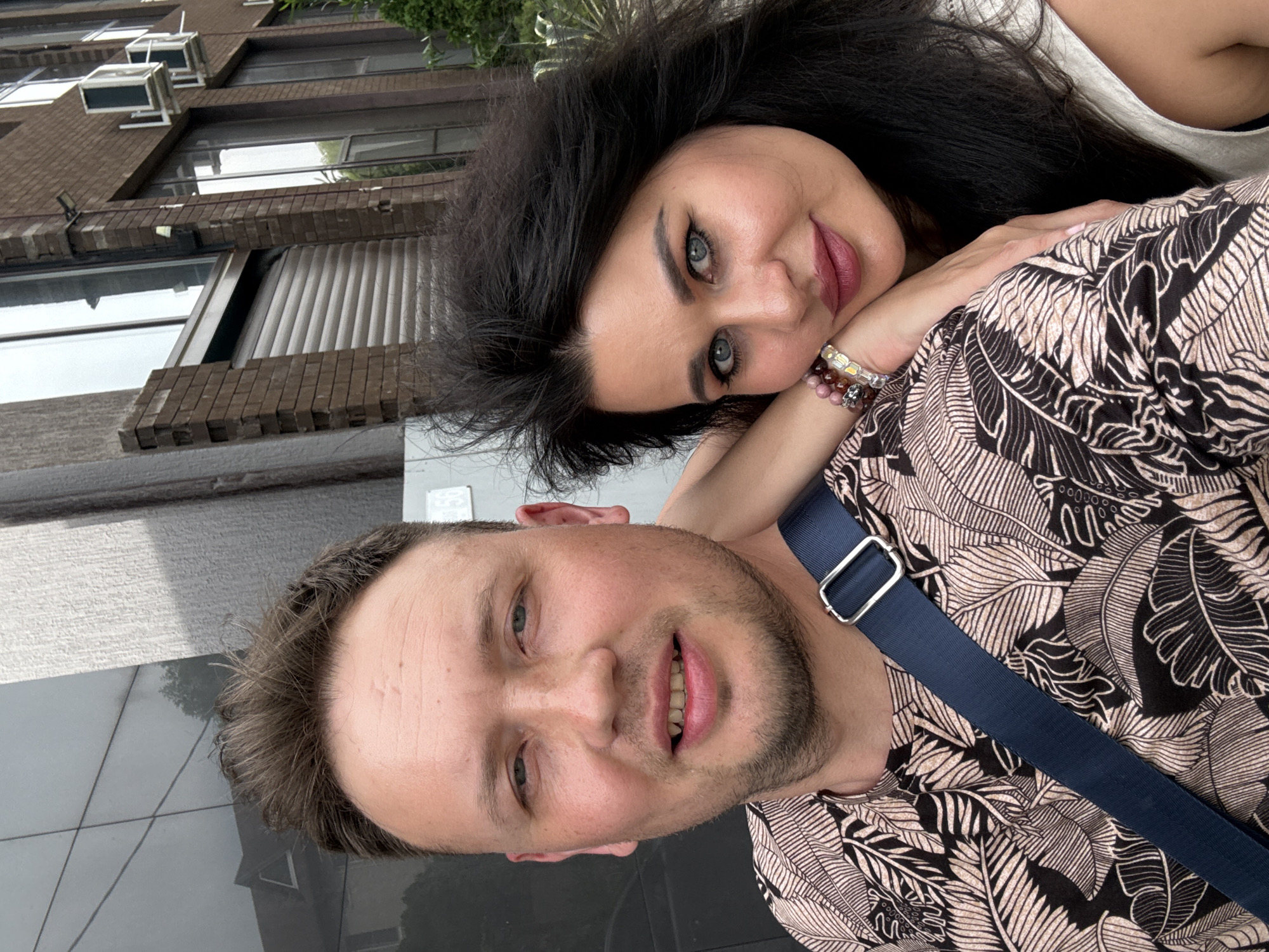 This is a close-up, outdoor, selfie-style photograph of a man and a woman, likely a couple, in the city of Sochi, Russia. The man, on the left, has short, light brown hair, fair skin, and is looking directly at the camera with a slight smile. He is wearing a dark t-shirt with a tropical leaf print and a dark blue strap across his chest, possibly from a bag. The woman, on the right, has long, dark, wavy hair and is leaning her head on the man's shoulder, looking towards the camera. She has fair skin, dark eyebrows, and is wearing makeup, including dark eyeliner and lipstick. She is wearing several bracelets on her left wrist. The background consists of a building with a brick facade, large windows, and air conditioning units, suggesting an urban commercial or residential area. The overall lighting suggests it is daytime.
