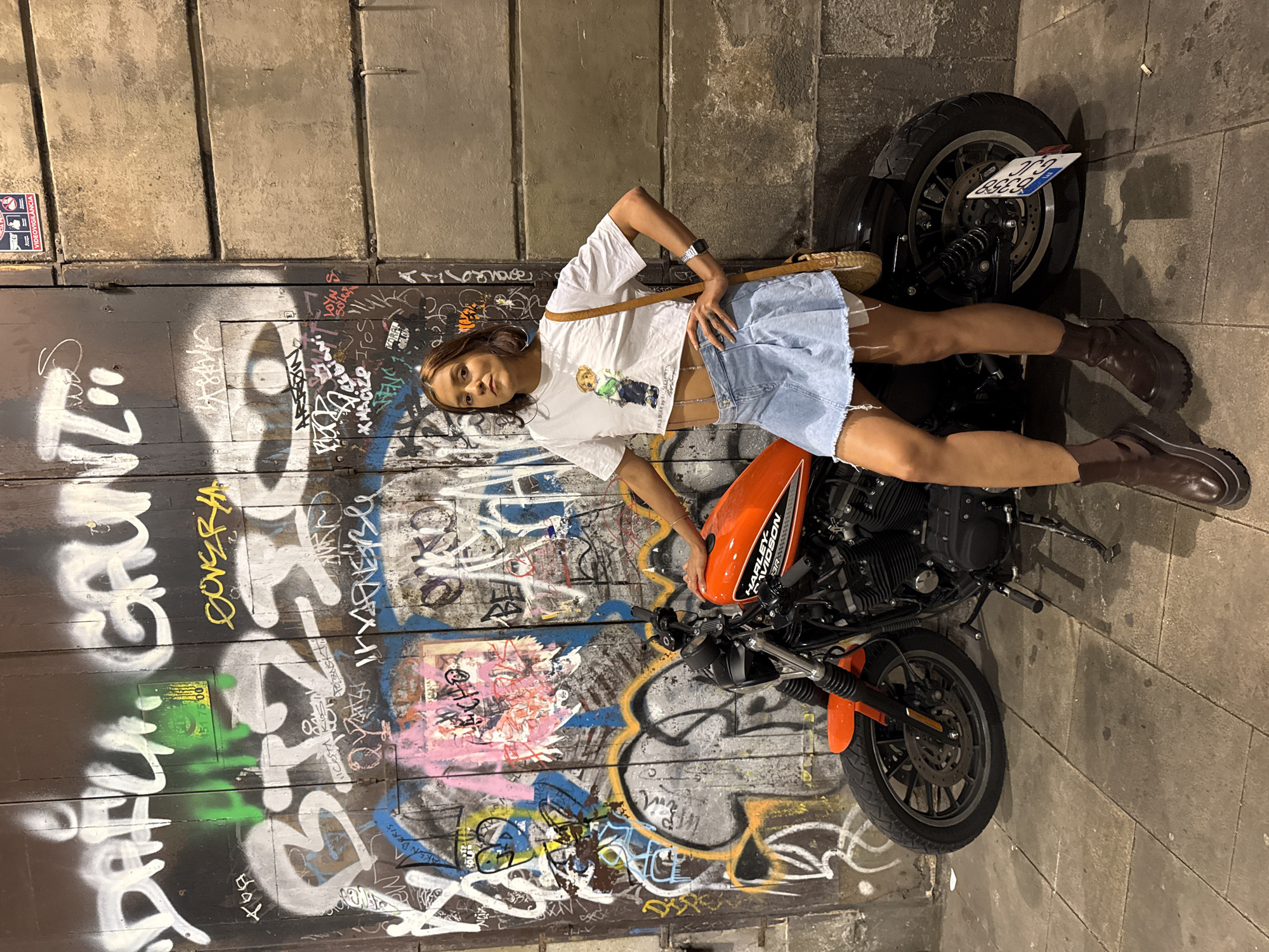 A young woman stands next to a motorcycle in front of a graffiti-covered wooden door in Barcelona, Spain. She has dark hair and is wearing a white crop top with a teddy bear graphic, a light blue denim skirt, and dark brown boots. She is posing with her hand on her hip and the other resting on the motorcycle. The motorcycle is orange with black accents and has the Harley-Davidson logo on its fuel tank. The wooden door is covered in various graffiti tags and drawings, with some paint peeling off. The ground is paved with stone tiles. To the right of the door, a section of a concrete building is visible.