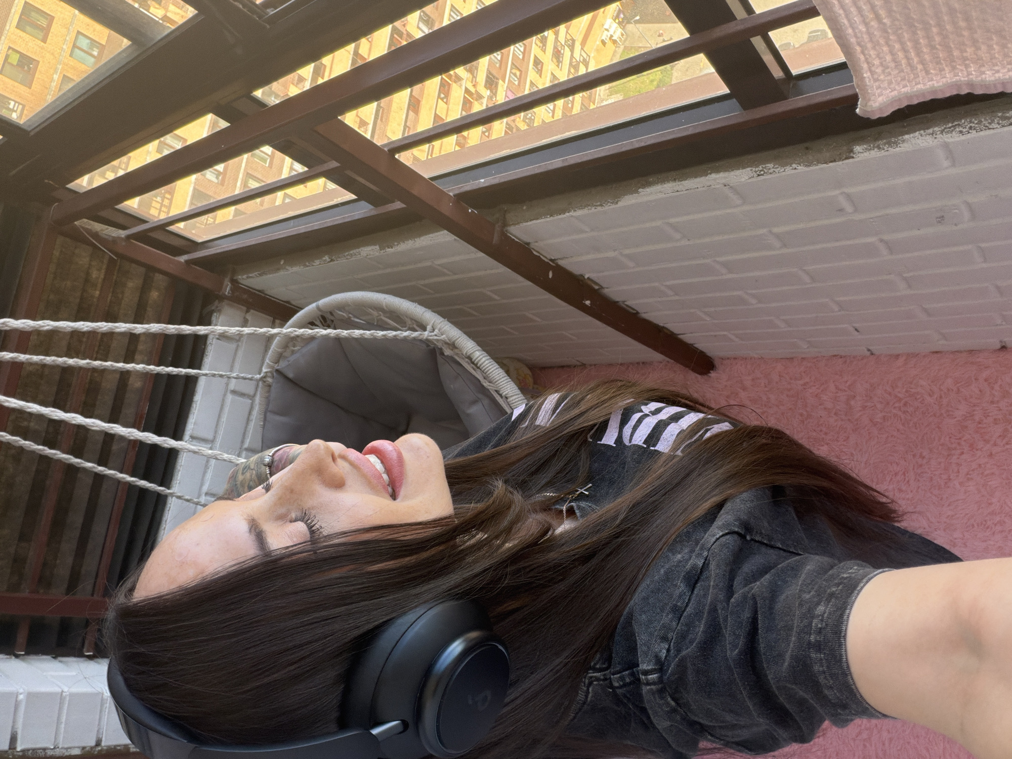 A young woman with long, dark brown hair is lying on a pink furry surface. She is wearing black headphones and a dark, acid-wash denim jacket. A white hammock-style chair is visible in the background to her left, alongside a white brick wall. Above, a brown metal structure frames a view of buildings in the distance. The specific location of Novosaratovka, Russia, cannot be confirmed from the image alone.