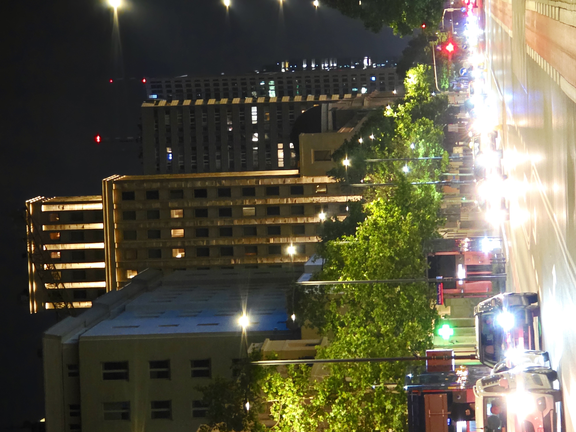 The image captures a night scene on a city street in Warsaw, Poland, likely near the Palace of Culture and Science given the prominent architecture. The foreground shows a road with several cars driving and stopped, their headlights illuminating the asphalt. A white SUV and a darker colored SUV are most visible. On the left side of the street, a building with several windows is visible, its façade illuminated by streetlights. Trees with lush green foliage line the street, their leaves catching the light. In the background, several tall buildings rise into the night sky. One of these buildings is a distinctive art deco style structure, brightly lit with vertical lines of light. Further back, other modern high-rise buildings are visible, some with illuminated windows and red lights atop antennas. A glowing green cross, likely indicating a pharmacy, is visible on the right side of the street, along with street signs and traffic signals, including a red traffic light. The overall atmosphere is one of a bustling urban environment at night.