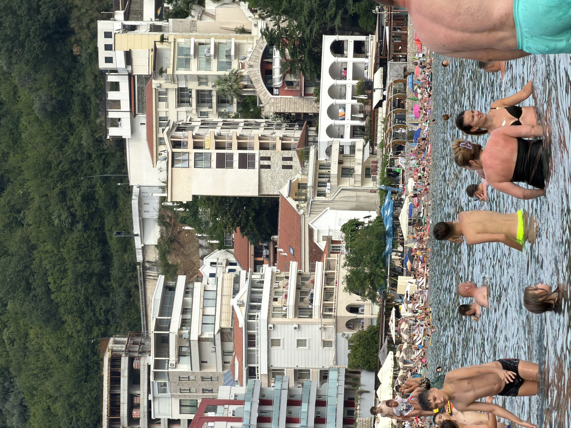 This image depicts a bustling beach scene in Rafailovići, Montenegro. The primary subjects are numerous people enjoying a day at the beach and in the water. They are mostly clothed in swimwear, with some visible in the water and others on the sandy shore.
The scene is set against a backdrop of densely vegetated hills and a resort-like area characterized by multi-story buildings with balconies and awnings. The architecture appears modern, with clean lines and light-colored facades. There are numerous beach umbrellas providing shade for people relaxing on the sand. The water is calm and appears to be the Adriatic Sea.
The activity taking place is recreational. People are swimming, wading in the water, and relaxing on the beach. The atmosphere seems lively and casual, typical of a summer beach day.
Notable details include the clear daylight, suggesting it is daytime, likely mid-morning to late afternoon, given the sun's position and the general brightness of the scene. The weather appears to be warm and sunny, conducive to beach activities. The presence of a resort area and the architectural style hint at a popular tourist destination. Some people appear to be in good spirits, engaged in conversation and enjoying the water.
There is some text visible on a yellow sign on one of the buildings which reads "FOR SALE". Other text is present on awnings and umbrellas, but it is too small and indistinct to read.