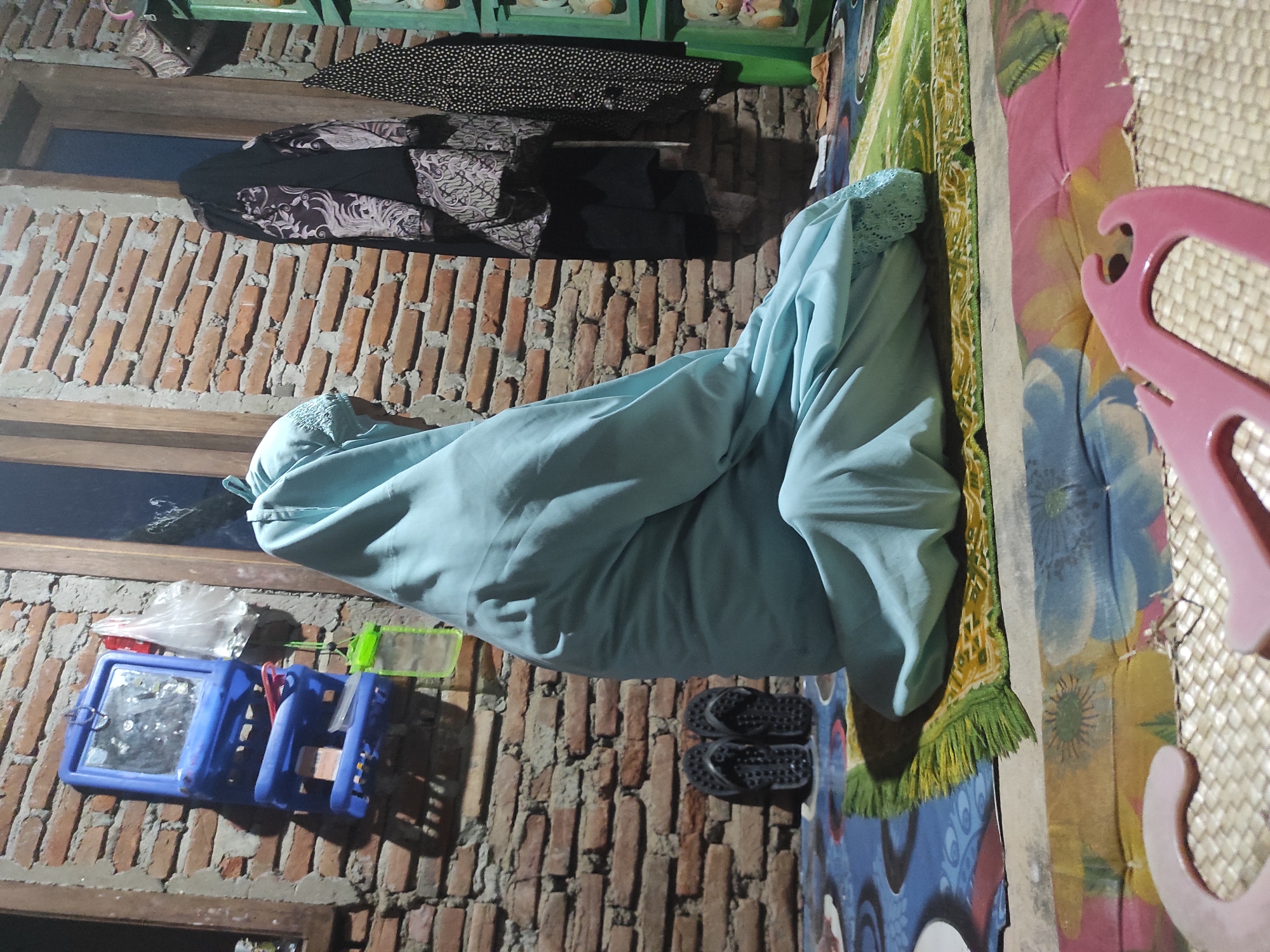 A person wearing a light blue robe is kneeling on a prayer rug in a room with brick walls. A blue plastic organizer with a mirror and other items are on the wall. Clothes hang above. Sandals are near the prayer rug. The location is Lhokseumawe, Indonesia.