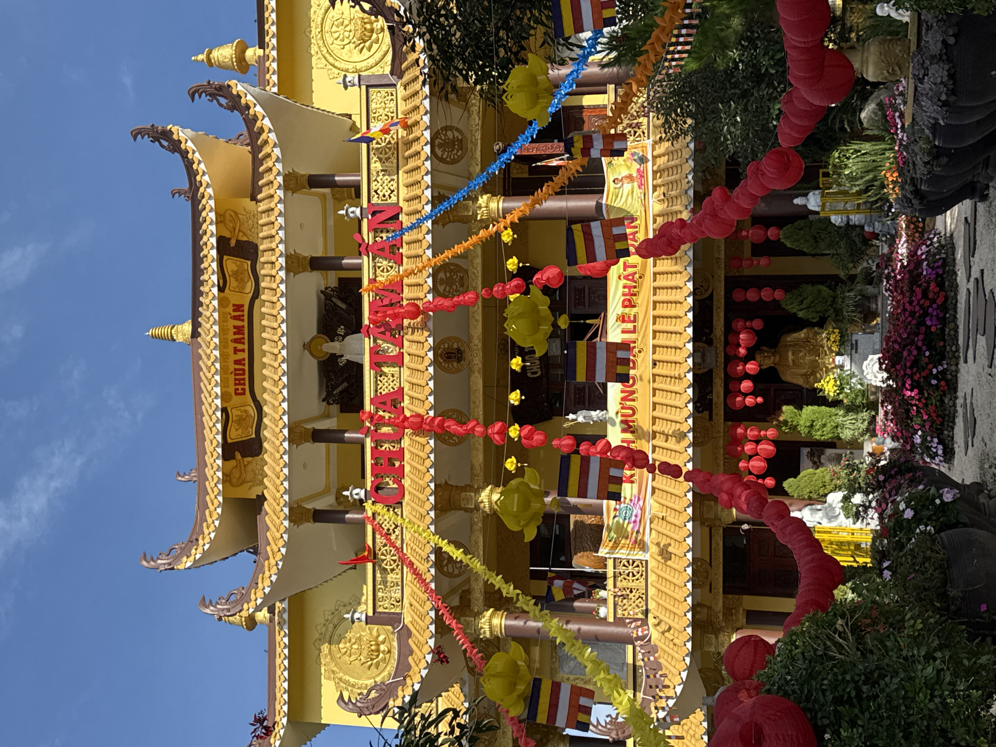 The image shows a vibrant, yellow Buddhist temple adorned with red and yellow streamers and flags. The temple features traditional Vietnamese architecture with curved roofs and ornate carvings. A large golden Buddha statue is visible in the foreground, surrounded by lush greenery and colorful flowers. The sky is clear and blue, suggesting a sunny day. The temple is likely the Chua Tam An pagoda, a well-known landmark in Da Lat, Vietnam, famous for its distinctive architecture and religious significance. The overall scene conveys a sense of peace and spiritual serenity, characteristic of Buddhist temples in Vietnam.