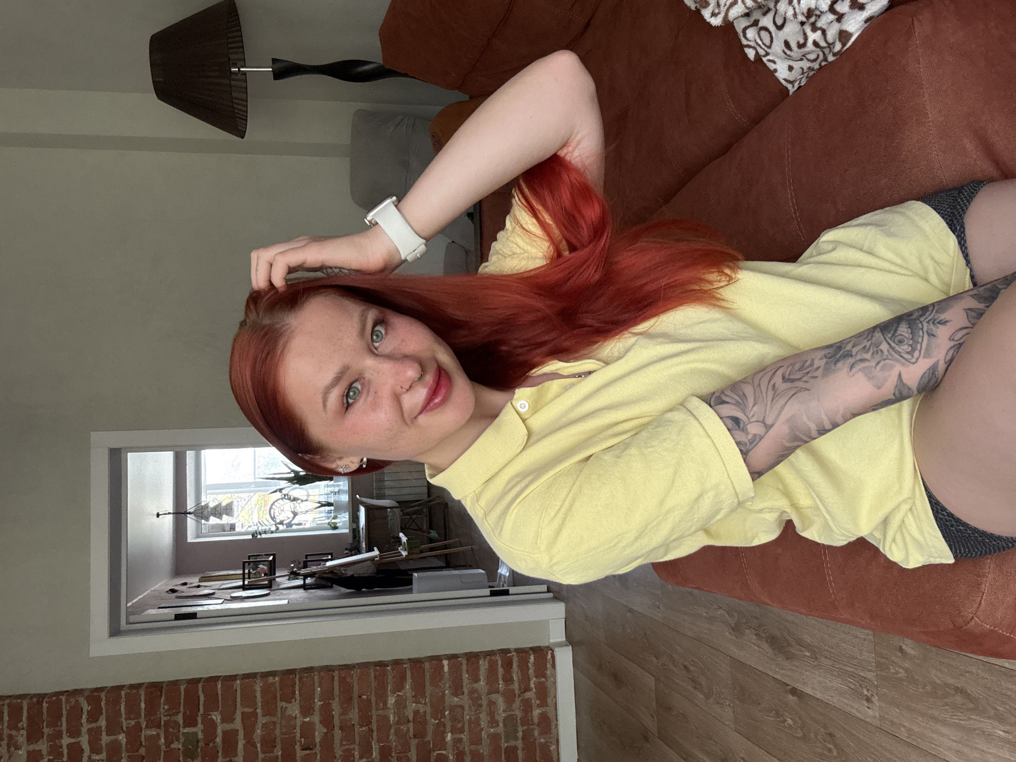 A fair-skinned woman with freckles and red hair is lying on a brown couch, wearing a yellow polo shirt and what appears to be dark-colored shorts or underwear. She is smiling at the camera with her right arm bent and hand touching her head, and her left arm, which is tattooed, is resting on her lap. She is wearing a white smartwatch.
In the background, there is a lamp, and a glimpse of a display case inside a room with an exterior brick wall. The location cannot be determined as Rostov-on-Don, Russia, based on the given image.
