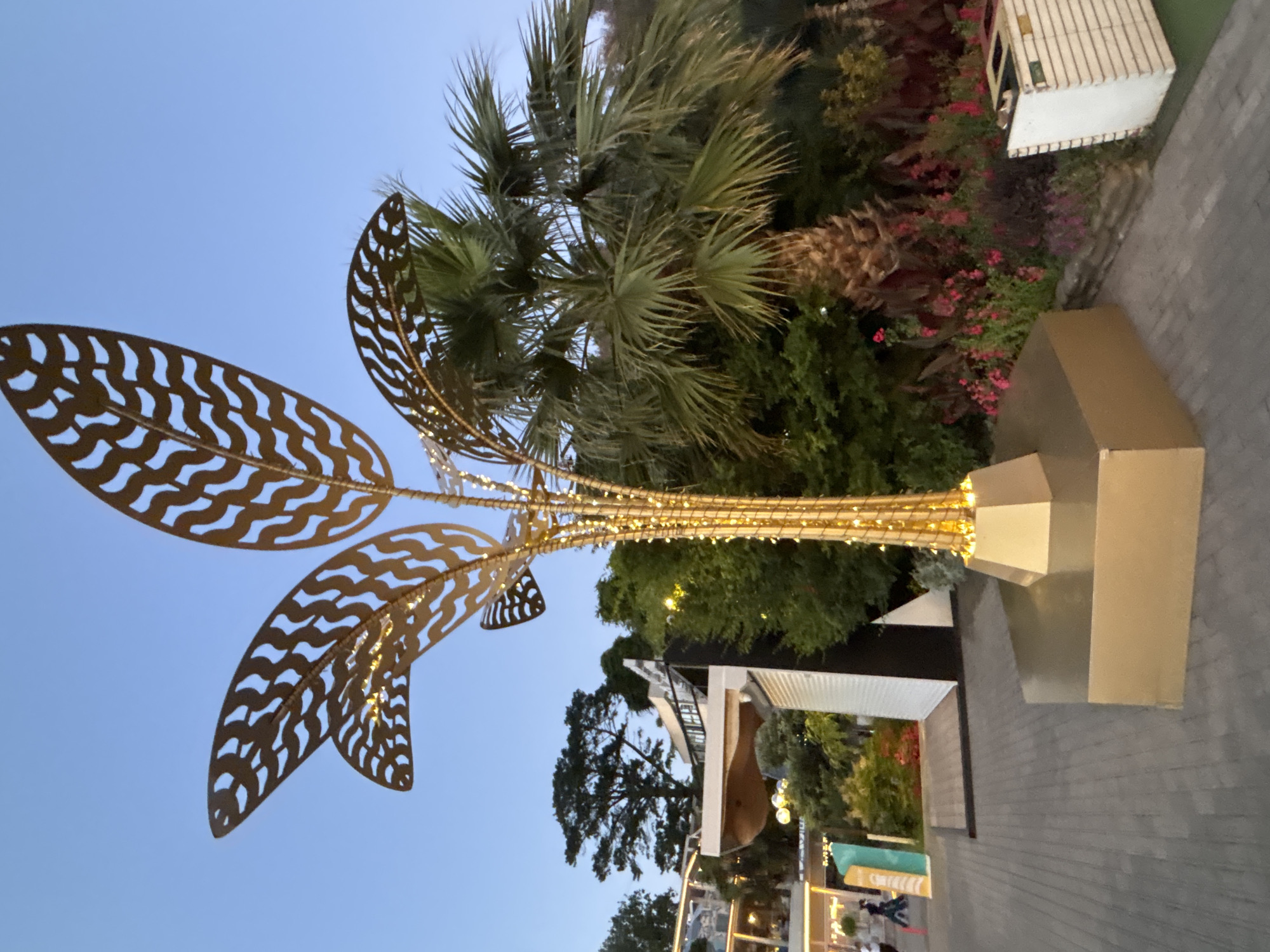 An outdoor decorative structure resembling a golden palm tree adorned with small, warm-toned lights stands on a paved walkway. The sculpture features intricate, leaf-like cutouts and is illuminated by the embedded lights. Behind the sculpture, a mature palm tree with fronds is visible, alongside lush green foliage and some vibrant pink flowers. In the background, a multi-story building with a modern design, featuring balconies and a unique curved roof, is partially obscured by trees. The sky above is a clear, twilight blue. The scene appears to be an outdoor public space, possibly a park or promenade, located in Sochi, Russia.