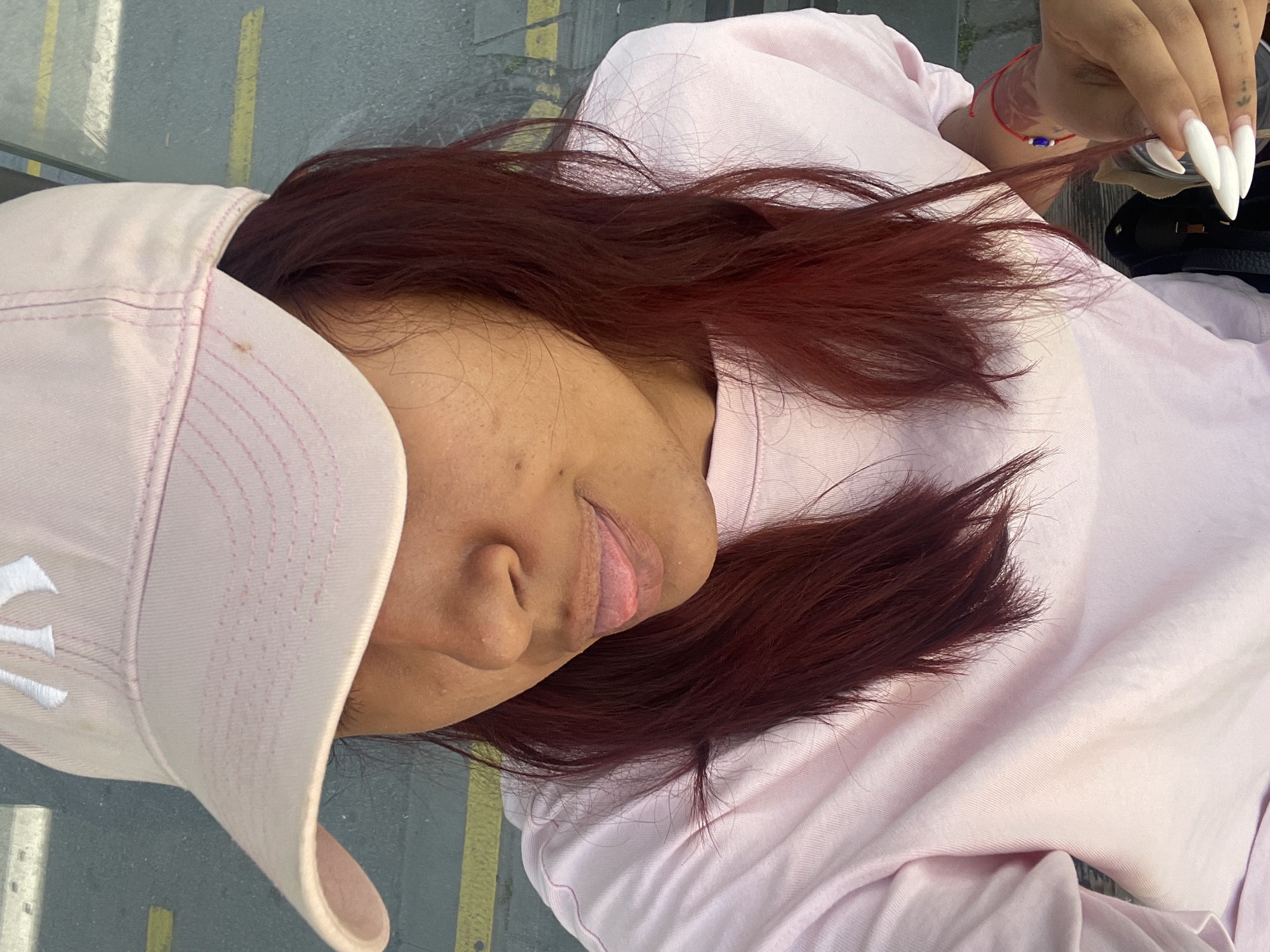 A young woman wearing a pink baseball cap and a pink t-shirt is pictured from a close-up, high-angle perspective. She is sticking her tongue out slightly. Her long, dark red hair is visible around her face and shoulders. She is wearing a red string bracelet on her left wrist. Her left hand is visible, with long, white pointed fingernails. She appears to be sitting outdoors, with a blurred background that suggests an urban environment with yellow lines on asphalt, possibly a street or sidewalk. The overall lighting suggests it is daytime. The specific location in Varna, Bulgaria, cannot be determined from the visual information alone.