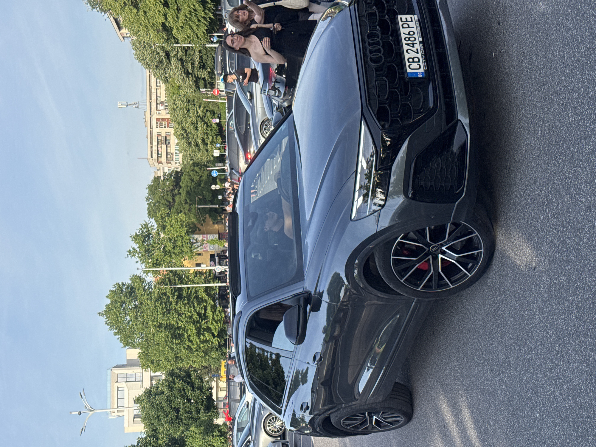The image captures a black Audi RS Q8 with the license plate "CB 2486 PE" parked on a street in Plovdiv, Bulgaria. There are two women standing near the front of the car, one in a long black dress and the other one behind her wearing a black top and black pants, possibly posing for a photo. Other vehicles are visible in the background, parked along the street, including what appears to be a silver SUV. A crowd of people is seen behind the cars on the left. There are trees lining the street, and buildings can be seen in the background. The sky is clear and blue. The street appears to be paved with asphalt.