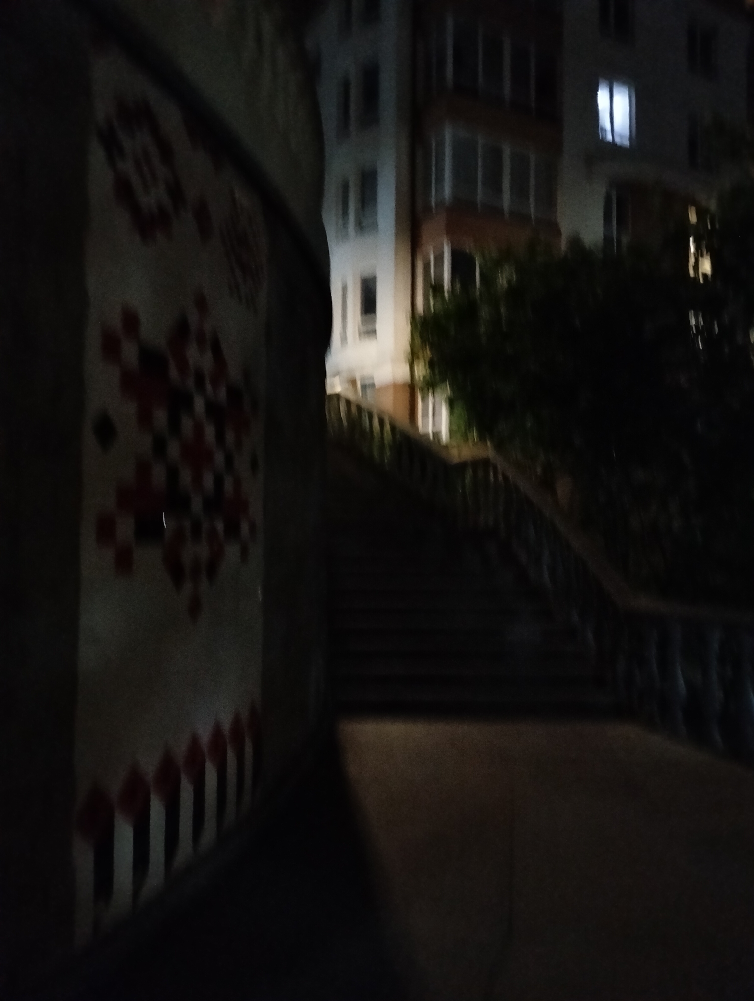 The image shows a set of outdoor stairs in Krykhivtsi, Ukraine, at night. To the left, there is a decorated curved wall with geometric patterns. In the background, a multi-story building is visible, with some windows illuminated. A tree partially obscures the view of the building. The stairs are dark, and the area around them is dimly lit.
