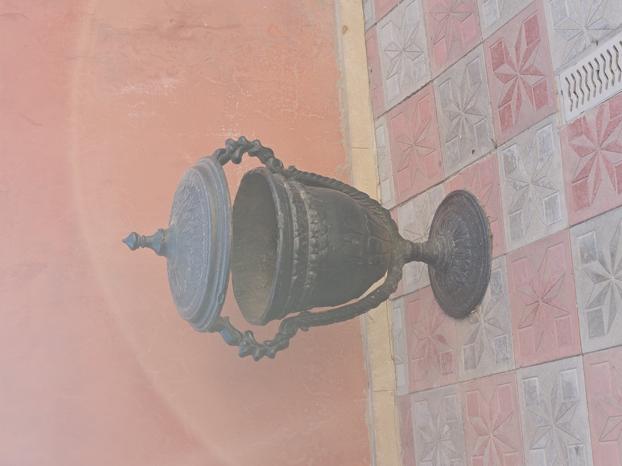 A black, ornate cast-iron trash receptacle stands on a patterned tile walkway in front of a textured pink wall. The trash receptacle has decorative rope-like handles and a decorative finial on its lifted lid. The walkway consists of square tiles with a starburst pattern in alternating shades of pink and grey. A metal drain grate is visible on the right side of the walkway. The scene is outdoors, possibly in a public area. The location is identified as Sumqayıt, Azerbaijan. There are no people or animals visible in the image.