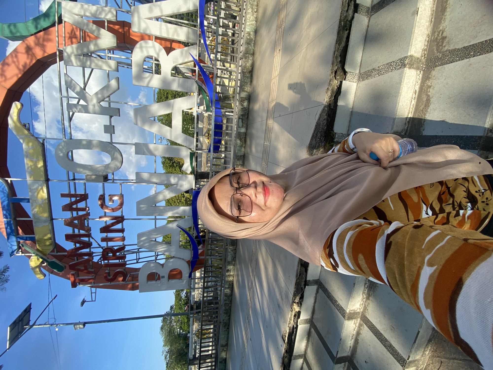 A woman is taking a selfie in front of a large landmark sign that reads "TAMAN SIRING 0 KM BANJARMASIN" in Banjarmasin, Indonesia. The sign is colorful and appears to be made of metal. The woman is wearing a beige hijab, glasses, and a long-sleeved shirt with a camouflage pattern in shades of brown, orange, and white. She is holding a bottle of water in her right hand. The background shows a blue sky with some clouds, trees, and a metal fence. The ground in front of the sign is paved with concrete and has a textured pattern. There are also some stairs leading up to the paved area. The lighting suggests it is daytime.