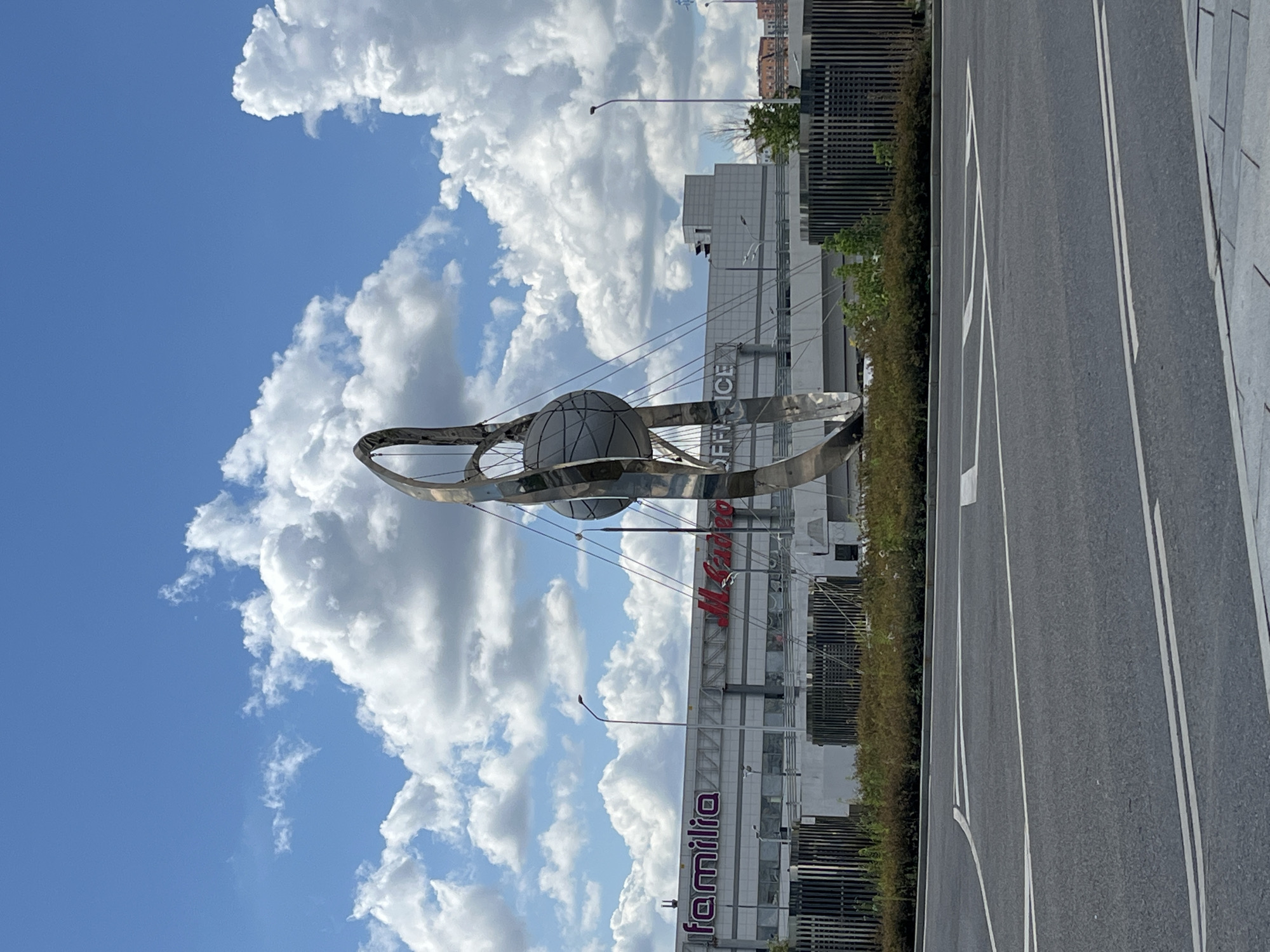 The image captures a modern outdoor sculpture in front of a large commercial building under a partly cloudy blue sky. The sculpture is metallic and abstract, featuring a spherical element suspended within curved metallic structures. The building behind it has "Familia" and "M. Video" branding visible, suggesting it is a shopping center or retail space. A road with white lane markings is in the foreground, indicating a street or parking area. The environment appears to be urban, possibly a commercial district in Moscow, Russia, given the context. There are no people visible in the frame.