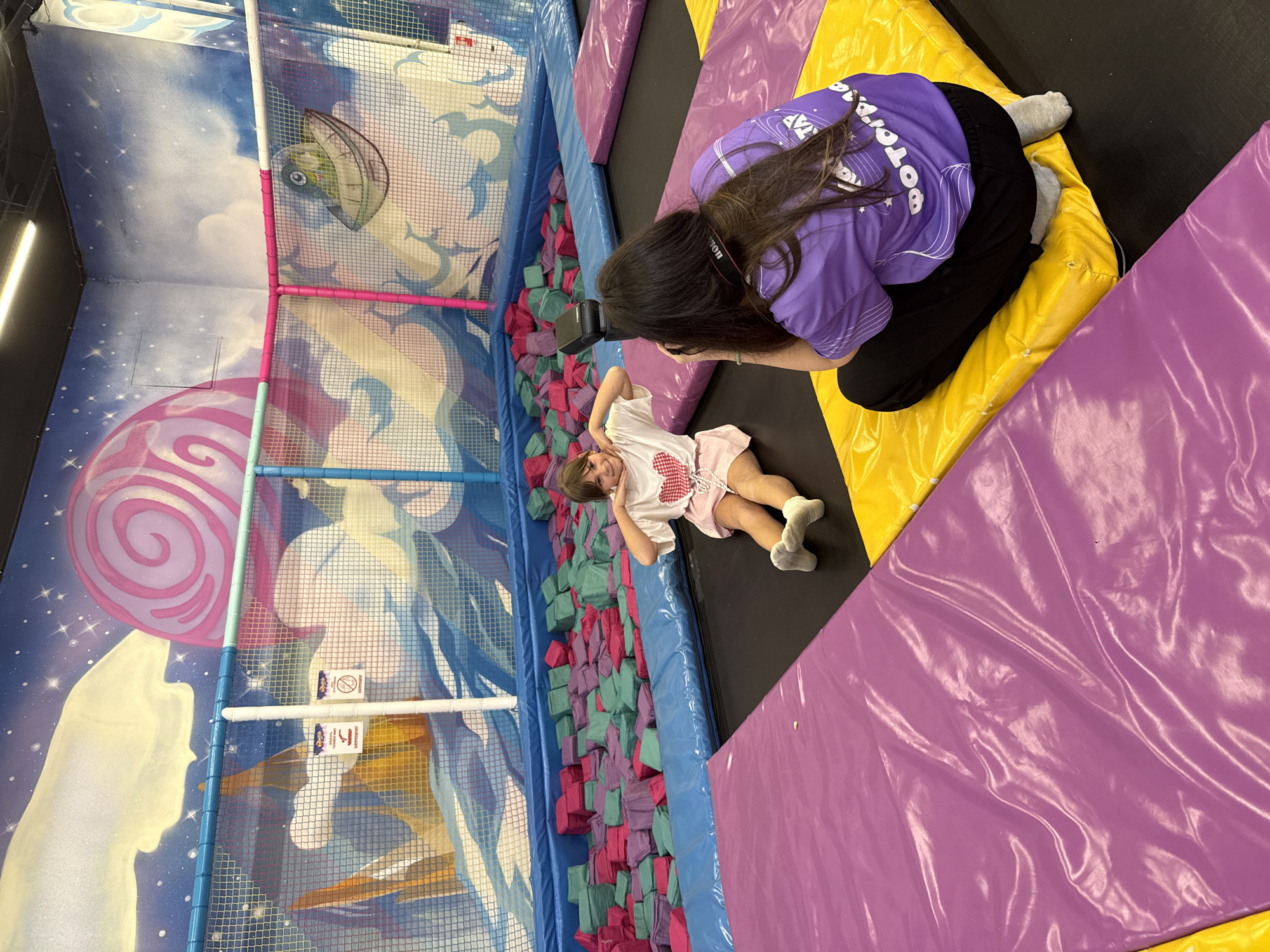 In Korobovo, Russia, a young girl with dark hair is posing for a photo on a trampoline. She is wearing a white t-shirt with a red heart design and a pink skirt. Her hands are on her cheeks in a playful gesture. Beside her, a person with long dark hair, wearing a purple t-shirt and black pants, is kneeling and taking a picture with a camera. They are both on a trampoline with purple and yellow mats. In the background, there is a foam pit filled with pink and green cubes and a mural on the wall depicting a fantasy scene with clouds, stars, and a large pink swirl.