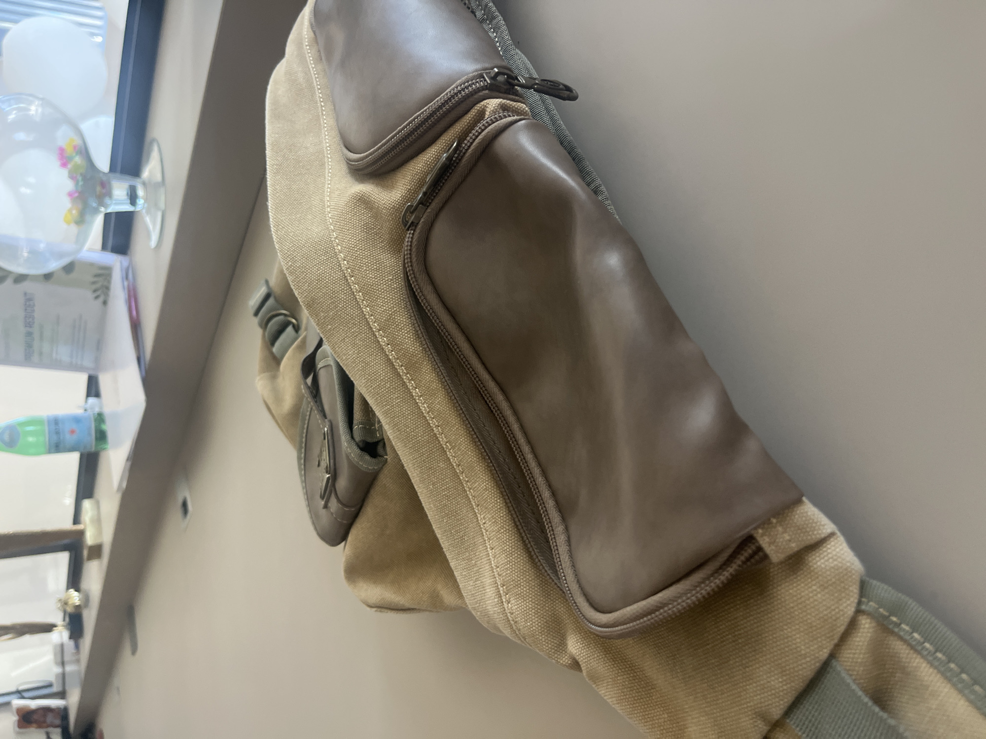 A tan canvas bag with brown leather accents is placed on a light-colored surface. The bag has multiple zippered compartments. In the background, a clear glass bowl filled with colorful small objects, possibly beads or marbles, sits on a windowsill. A green bottle of San Pellegrino water is also visible on the windowsill, along with a framed picture and a trophy. The environment appears to be indoors, likely in a modern building, given the clean lines of the furniture and the window. This scene is located in Germasogeia Municipality, Cyprus.