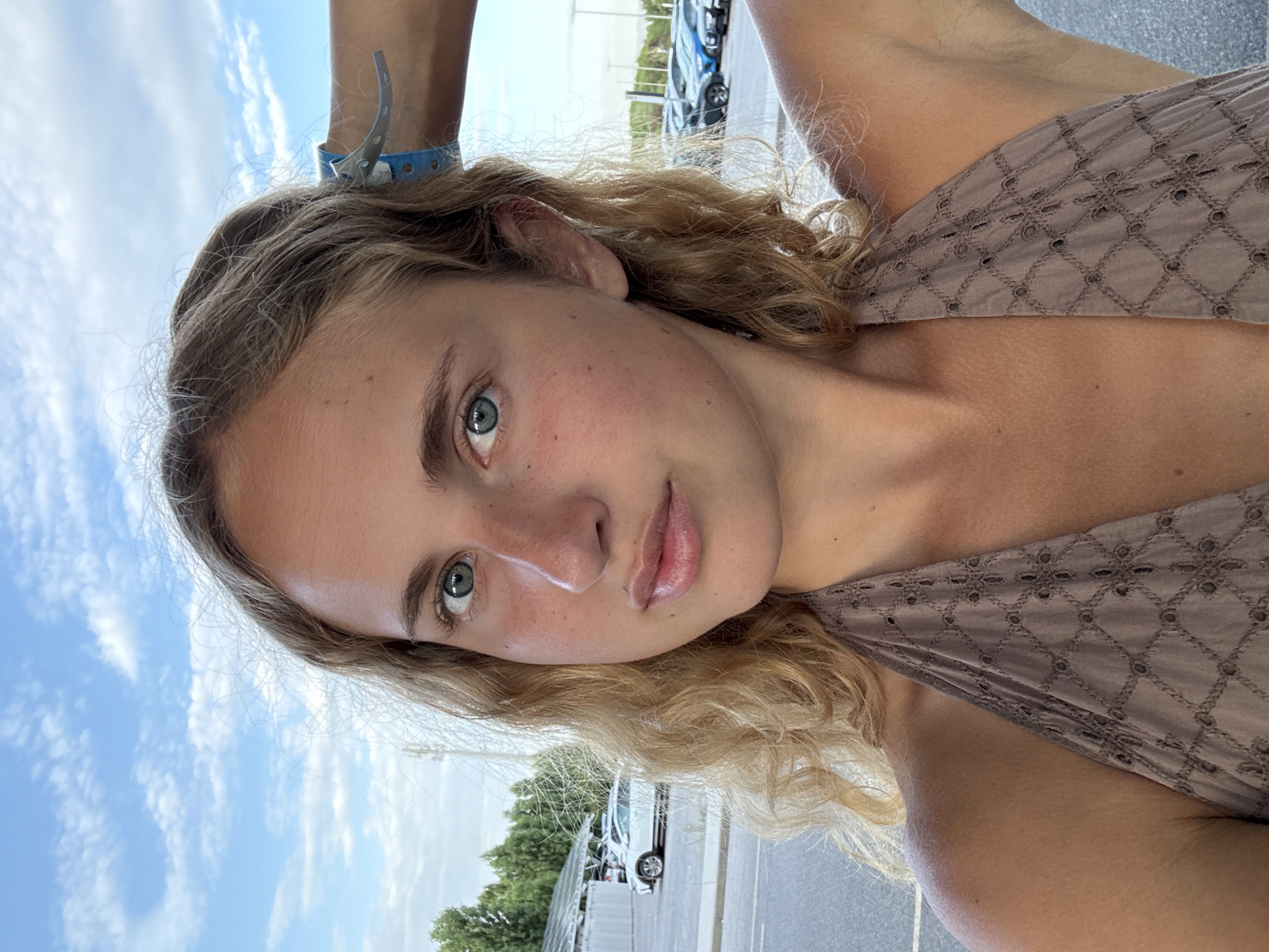 A young woman with blonde, curly hair and blue eyes looks at the camera in a selfie. She is wearing a brown halter top with an embroidered pattern. Her right hand is raised, resting on the back of her head, where a blue wristband is visible. The background shows a cloudy blue sky, trees, and a parking lot with several cars, suggesting an outdoor location. The scene appears to be a parking lot near a body of water, possibly a beach or coastline, as the sky and light suggest a seaside environment. While the exact location is not specified, the presence of cars and trees suggests an urban or suburban setting.