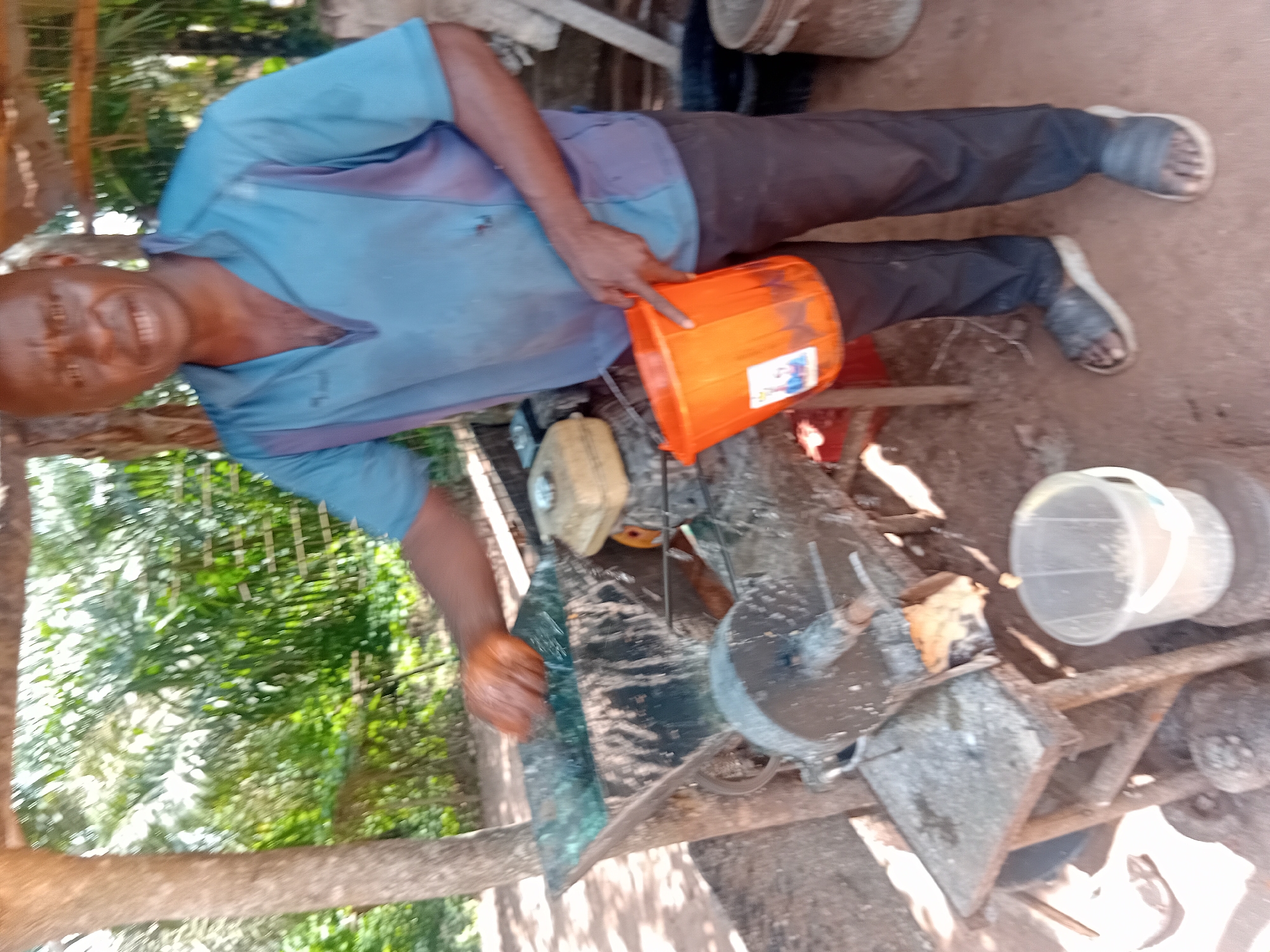 In Ejule, Nigeria, a man stands outdoors beside a manual processing machine, likely used for extracting oils or processing grains. He wears a light blue collared shirt and dark trousers, with dark sandals on his feet. In his right hand, he holds an orange plastic bucket, which has a label with an image on it. His left hand is near the feeding hopper of the machine, suggesting he is actively engaged in its operation. The machine itself is mounted on a wooden stand and appears to be made of metal, with a funnel-like opening at the top where materials are fed. Behind the man and the machine, a lush green environment with trees and foliage is visible, indicating a rural or semi-rural setting. The ground is covered in reddish-brown earth. The overall scene depicts a man engaged in manual labor within a natural, outdoor environment.
