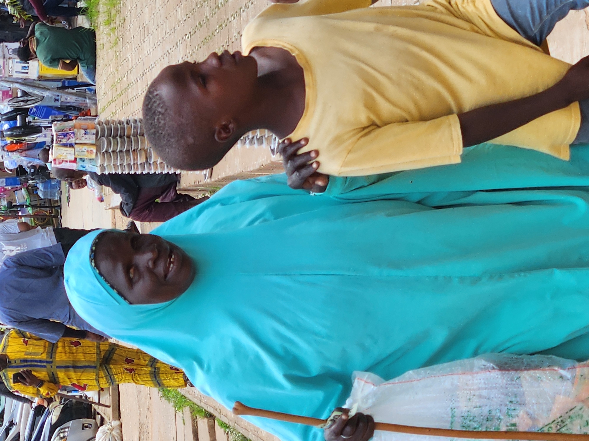 The image captures an outdoor scene in an urban setting, possibly a market. In the foreground, a woman wearing a vibrant turquoise headscarf and flowing robe is holding a wooden stick and a plastic bag. Her face, visible in the cropped image, shows a smile. To her right, a young boy in a yellow t-shirt and blue jeans stands with his arm around her shoulder. His expression is directed towards the right. In the background, other individuals are visible, some in traditional clothing, and a stall displaying various items including bottled water and packaged goods. The ground appears to be paved with bricks or concrete, with patches of grass and dirt. Cars are parked in the background, suggesting a busy thoroughfare or parking area.