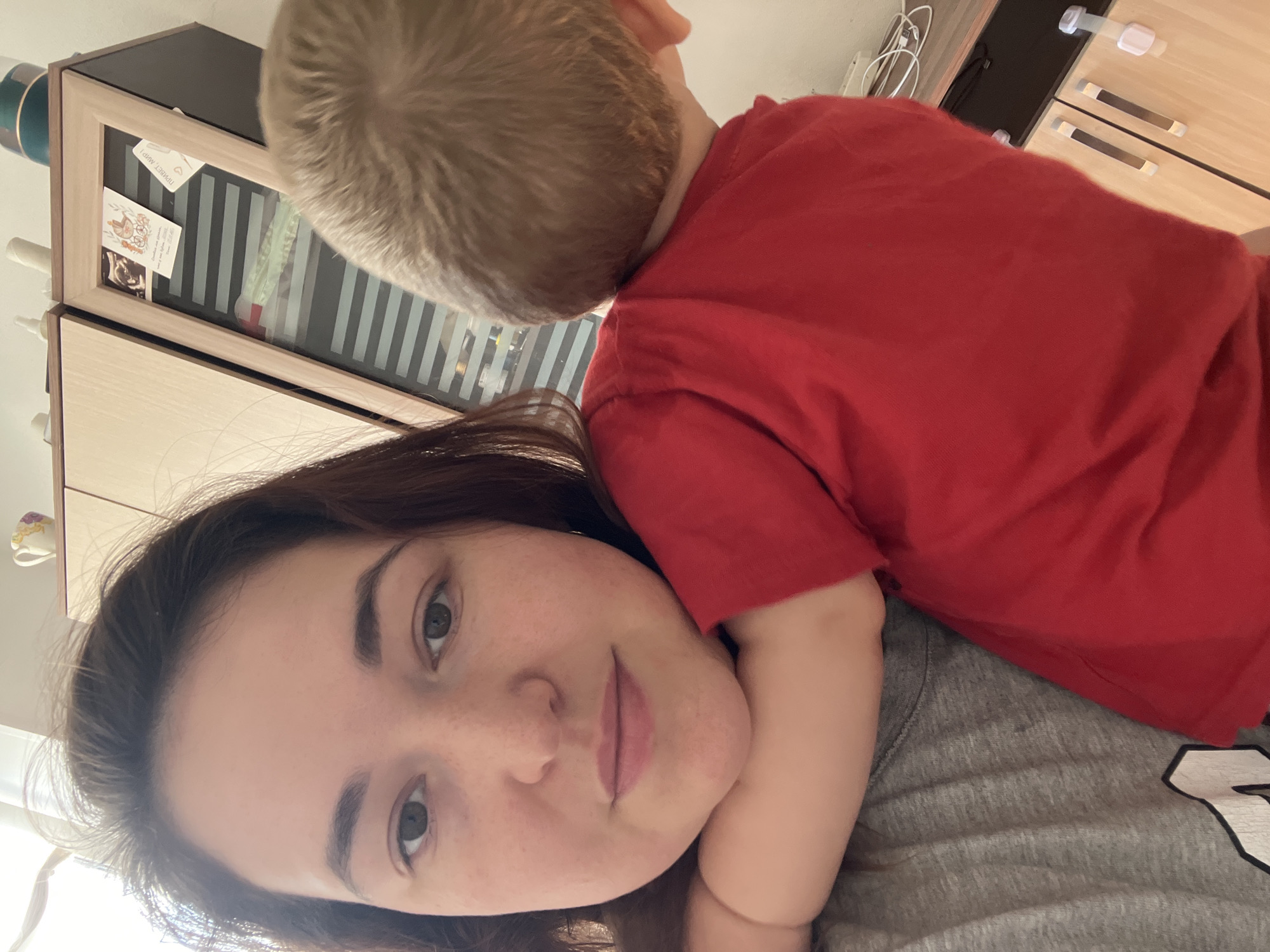 The image shows a close-up, selfie-style shot of a woman and a young child. The woman, with brown hair, is looking directly at the camera with a gentle expression. She is wearing a grey t-shirt. Resting on her shoulder and head is a young child, whose back and head are visible. The child is wearing a red t-shirt. The background appears to be an indoor setting, possibly a bedroom or living room, with a wooden cabinet or dresser visible. One of the drawers has a glass front, revealing some items inside, including what appears to be postcards or photographs. The lighting suggests it is daytime. There is no discernible weather information. The image evokes a sense of intimacy and affection between the woman and the child. No visible text is clearly legible in the image.