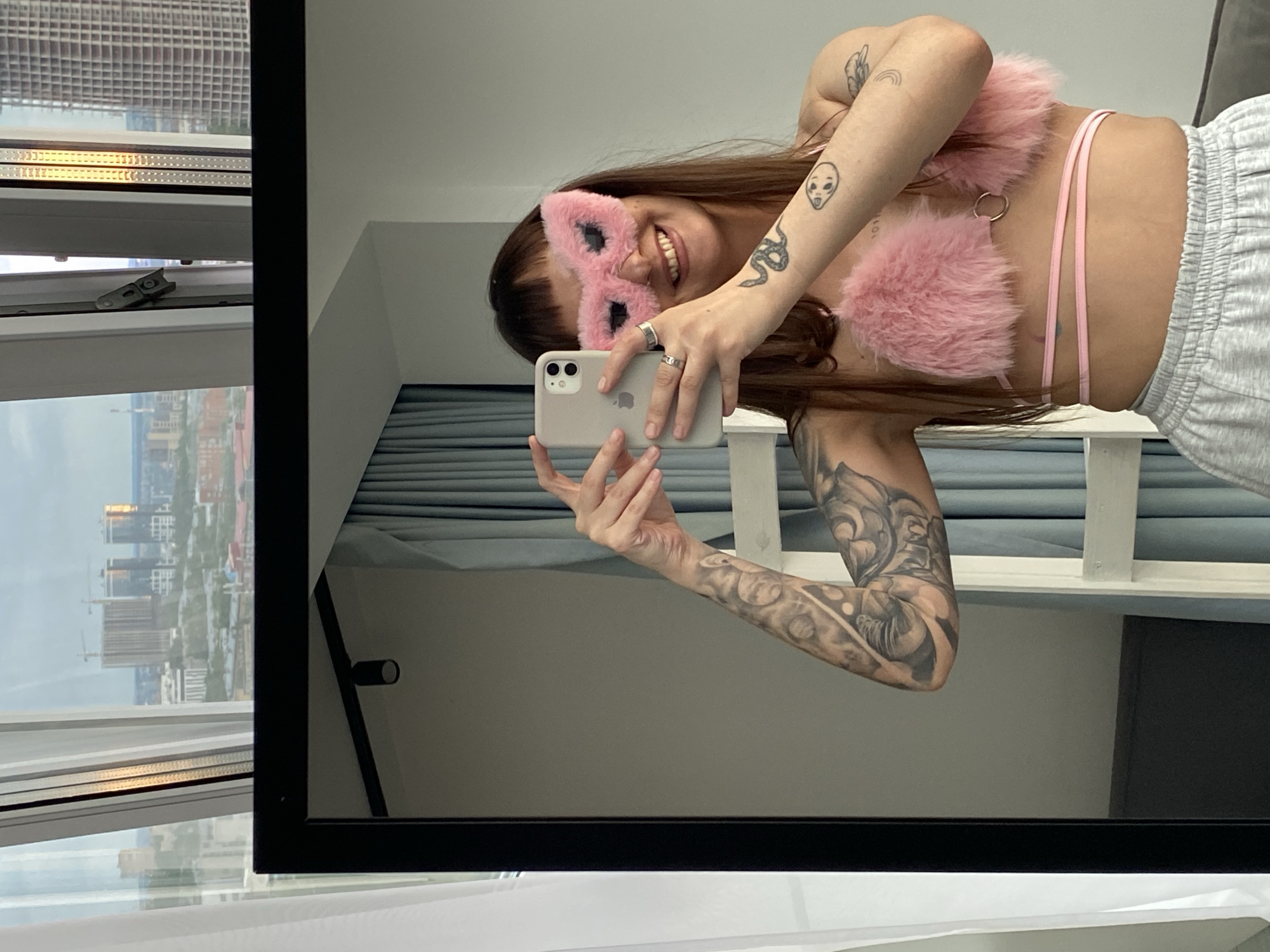 A young woman is taking a selfie in a mirror, holding her phone in her left hand. She is wearing a pink fluffy eye mask and a pink fluffy bralette with delicate straps. Her right arm is bent, and she is wearing rings on her fingers. Her left arm is visible, showing tattoos. She is smiling widely at her reflection, with her teeth showing. Her hair is long and brown, falling over her shoulder. She is wearing grey sweatpants.
The setting appears to be an indoor space with a large mirror reflecting the scene. Behind her, blue curtains are visible. To the left of the mirror, a window shows a cityscape with tall buildings and cranes, suggesting an urban environment. The view outside the window indicates it's daytime. The overall mood appears playful and relaxed. The image is cropped vertically from a larger image, with the mirror frame visible along the left and bottom edges.