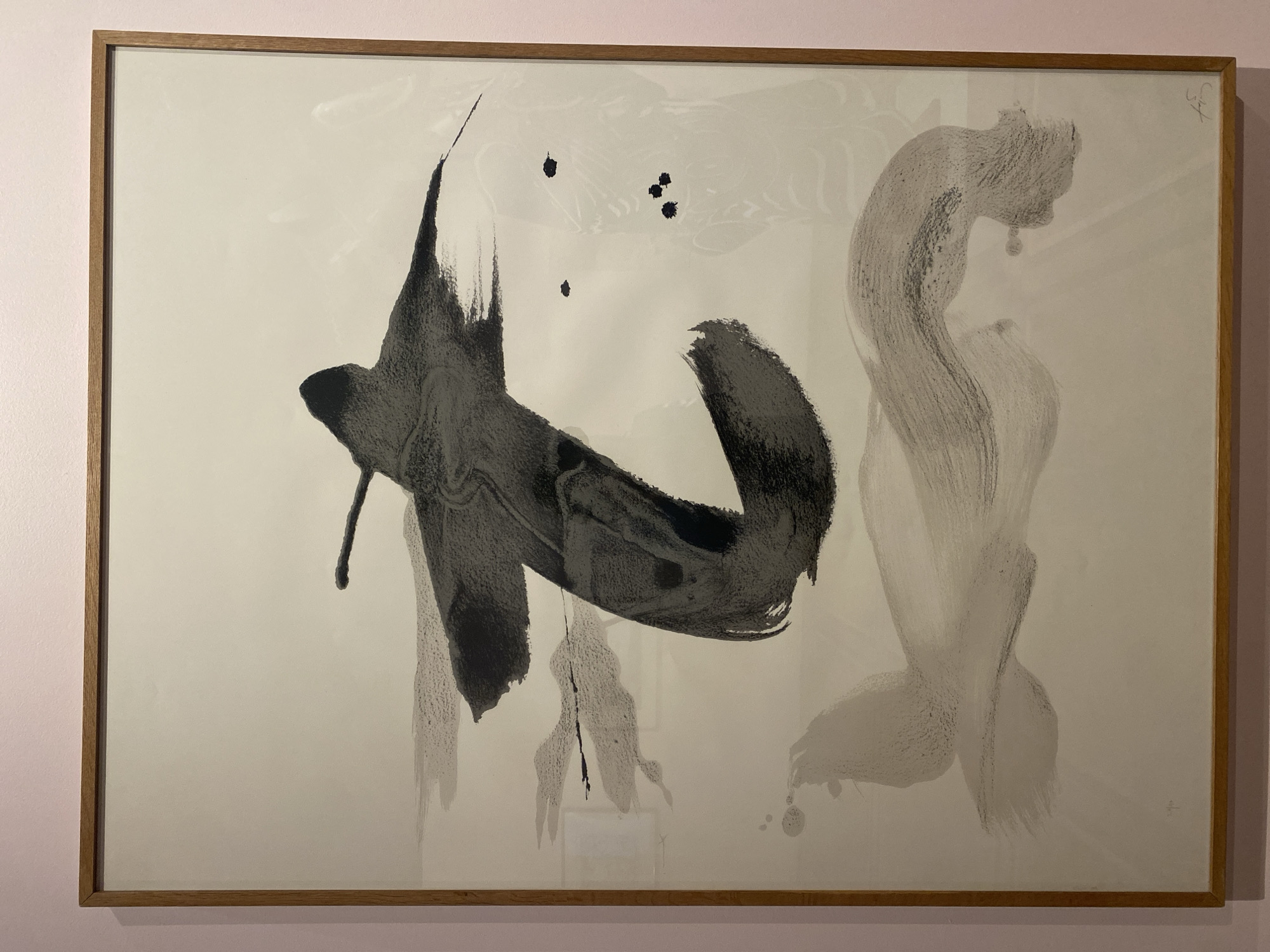 The image depicts a vertically oriented, framed abstract artwork displayed on a wall. The artwork features bold, calligraphic-style brushstrokes in black and various shades of gray on a white or off-white background. A large, dark gray to black brushstroke, resembling a stylized "T" or a character, dominates the upper-center of the composition. Lighter, sweeping gray brushstrokes are present in the mid-left and lower sections of the piece. Several small, circular black ink splatters are visible in the mid-right area. A faint mark or signature is located in the bottom-left corner, and a distinct signature is present in the bottom-right corner. The artwork is enclosed by a thin, light brown wooden frame and is protected by a reflective glass panel. The framed piece is mounted on a solid, pale pink wall. The scene is an indoor exhibition space or gallery located in Paris, France; specific architectural details or signage to identify the precise venue are not visible. No individuals are present within the frame.