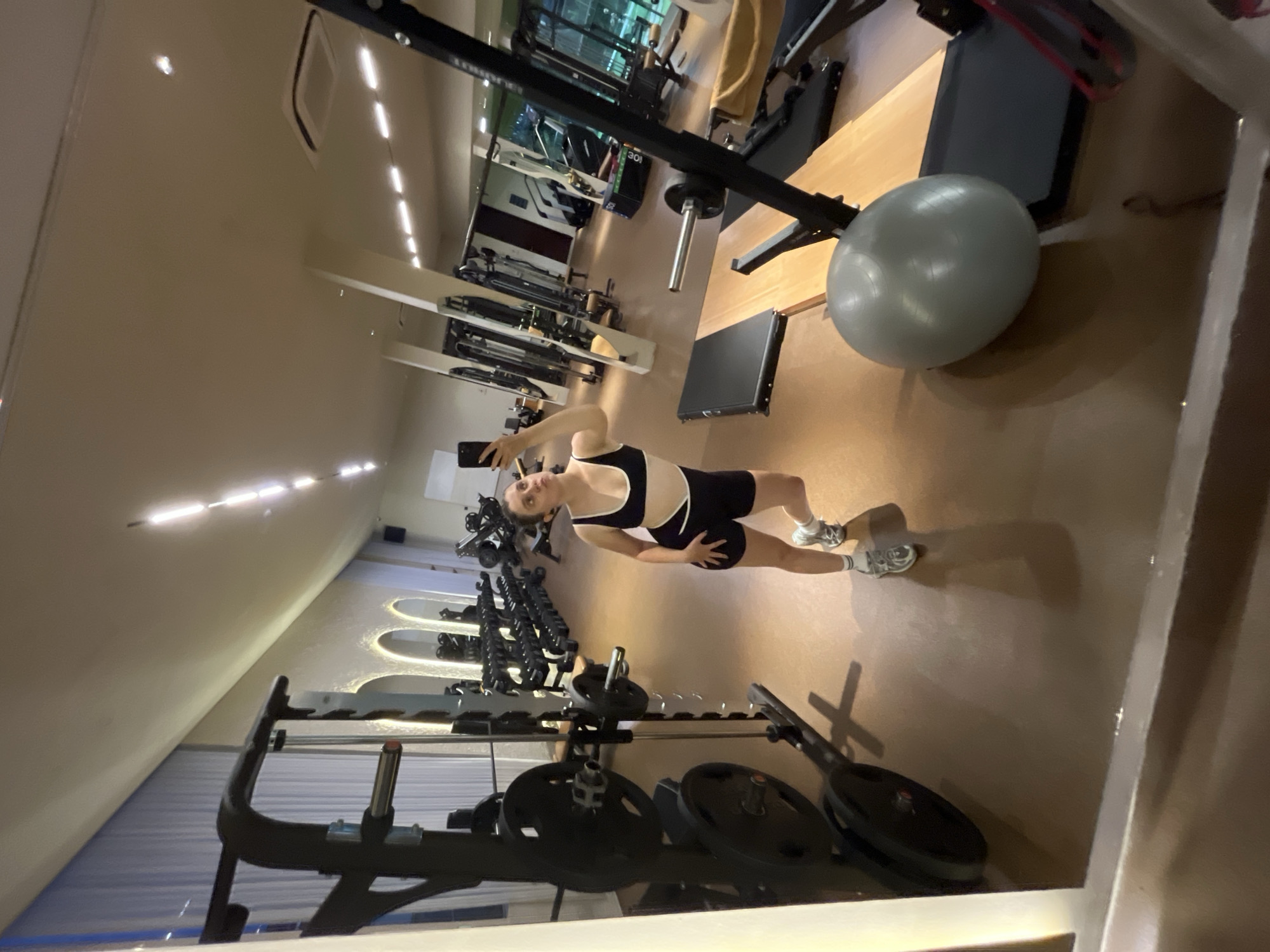 A woman stands in a gym, holding up her phone. The gym is filled with weightlifting equipment. The location is North Kuta, Indonesia.