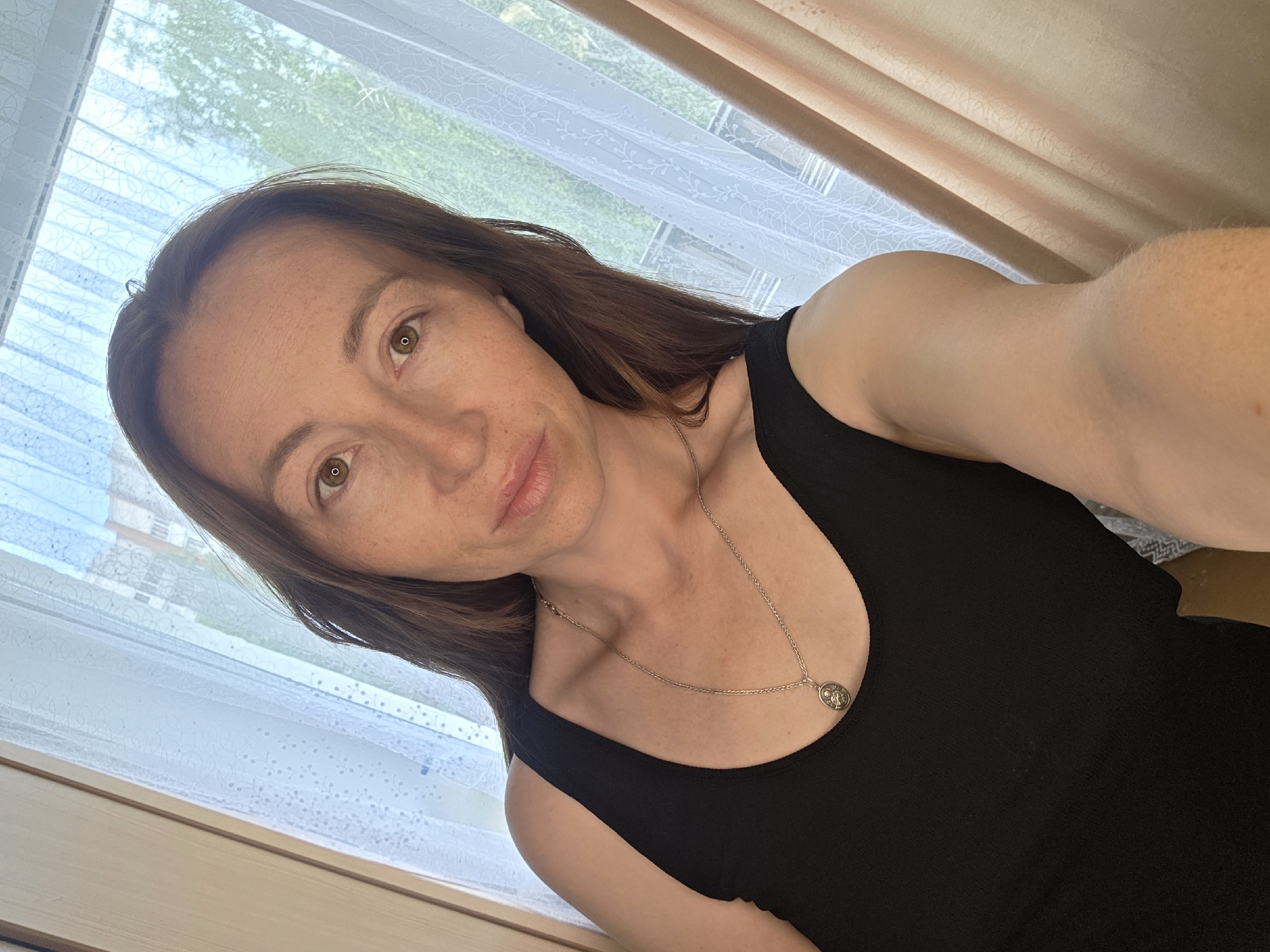 A woman is taking a selfie in a room with a window. She has long, brown hair and brown eyes and is wearing a black tank top and a silver necklace with a circular pendant. The background shows a window with sheer white curtains and partially drawn beige drapes, suggesting it is daytime. Through the curtains, it appears to be outdoors with trees visible, indicating a possibly residential or natural setting. The lighting suggests it could be a bright day, possibly morning or afternoon. The woman's expression is neutral, and she is looking directly at the camera. There is no visible text in the image.