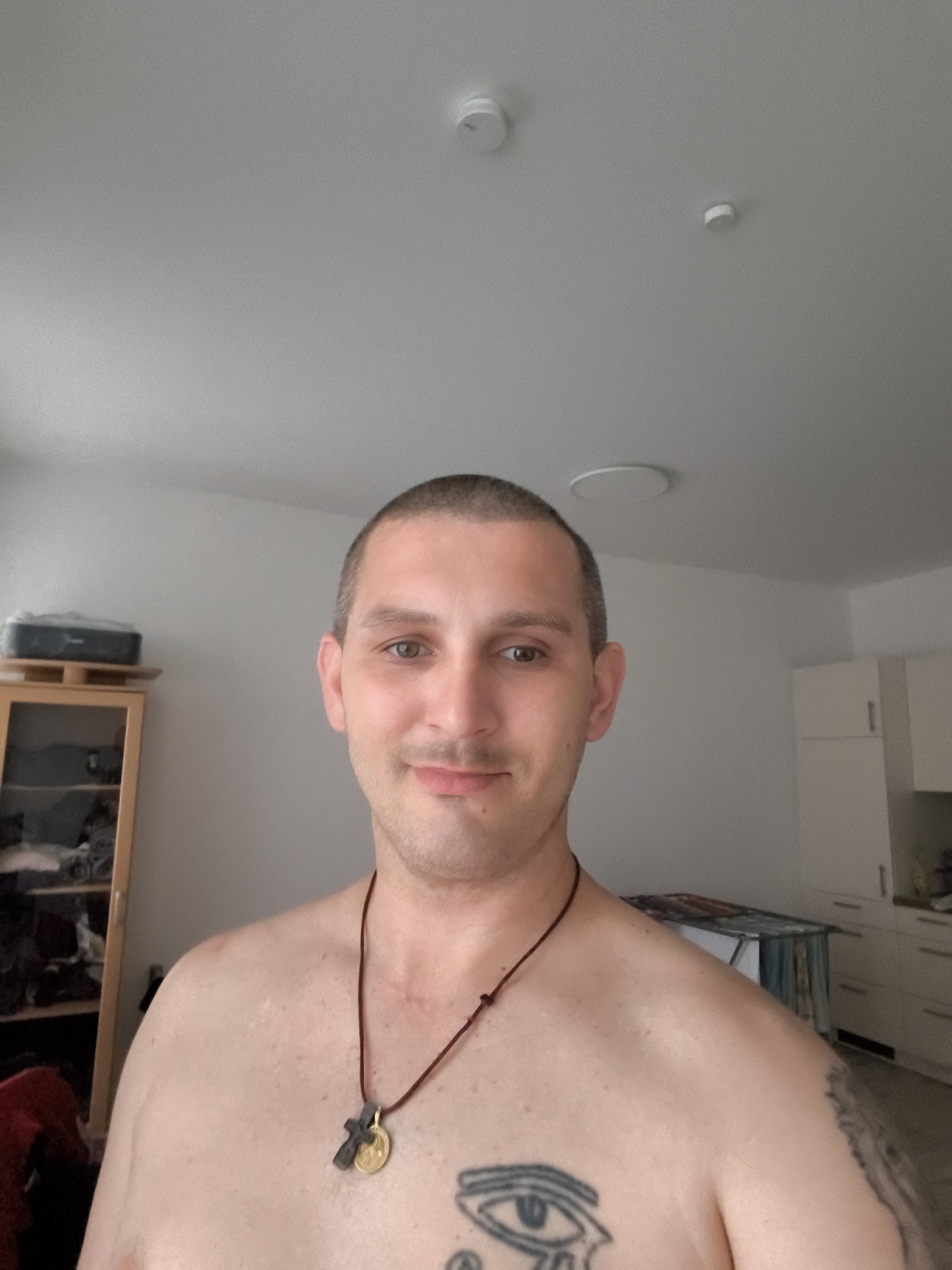 A male, appearing to be in his late 20s to early 30s, with short brown hair and brown eyes, is captured in a selfie-style photograph. He is shirtless, revealing a tattoo of an eye on his chest and another on his right arm. He wears a dark cord necklace with a pendant featuring a cross and a coin. He is indoors, in what appears to be a living space or bedroom. In the background, to the left, a tall wooden shelf unit with glass doors holds various items. To the right, white kitchen cabinets are visible, and a small table with a patterned cloth is in the mid-ground. The ceiling features two circular light fixtures. The overall lighting suggests an interior environment, possibly during daylight.
This image was taken in Wolgast, Germany.