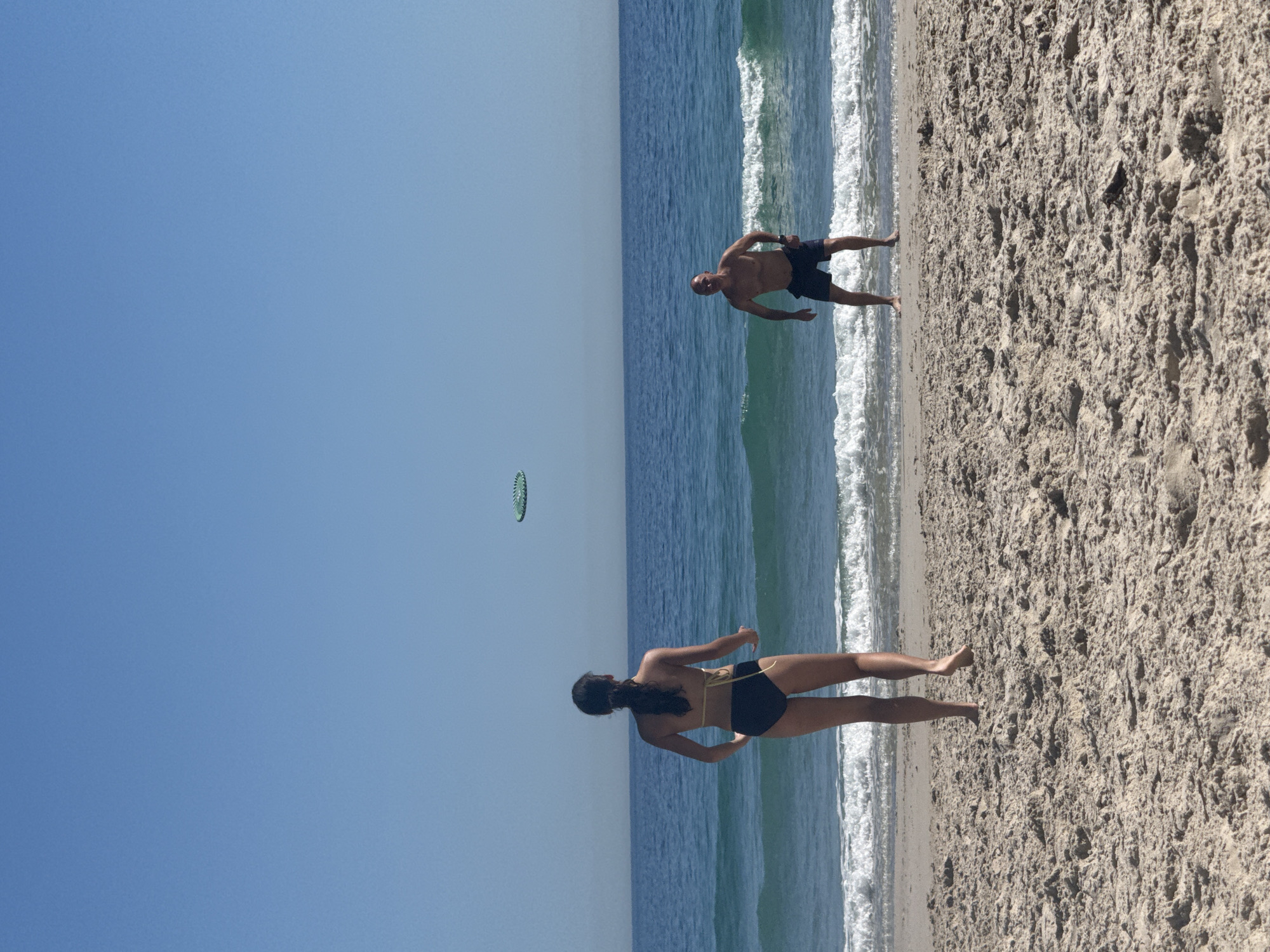 A woman and a man are on a beach near the ocean. The woman is wearing a black and tan swimsuit. The man is wearing black shorts. A green frisbee is in the air between them. The location could be Hout Bay, South Africa, given the sandy beach and the ocean.