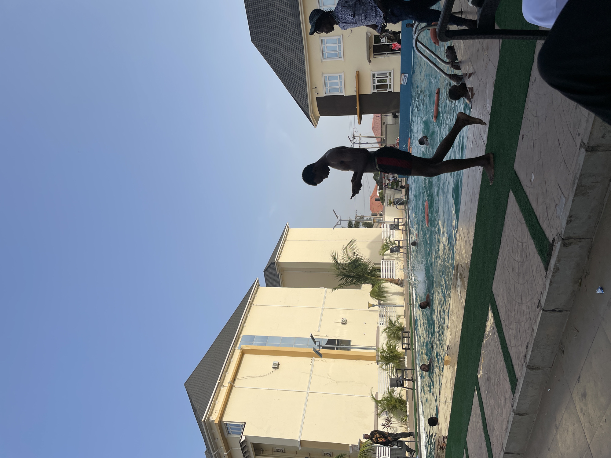 A man is jumping into an outdoor swimming pool. People are standing around the pool. Buildings and palm trees are visible in the background. The location is Barakin Lerawi, Nigeria.