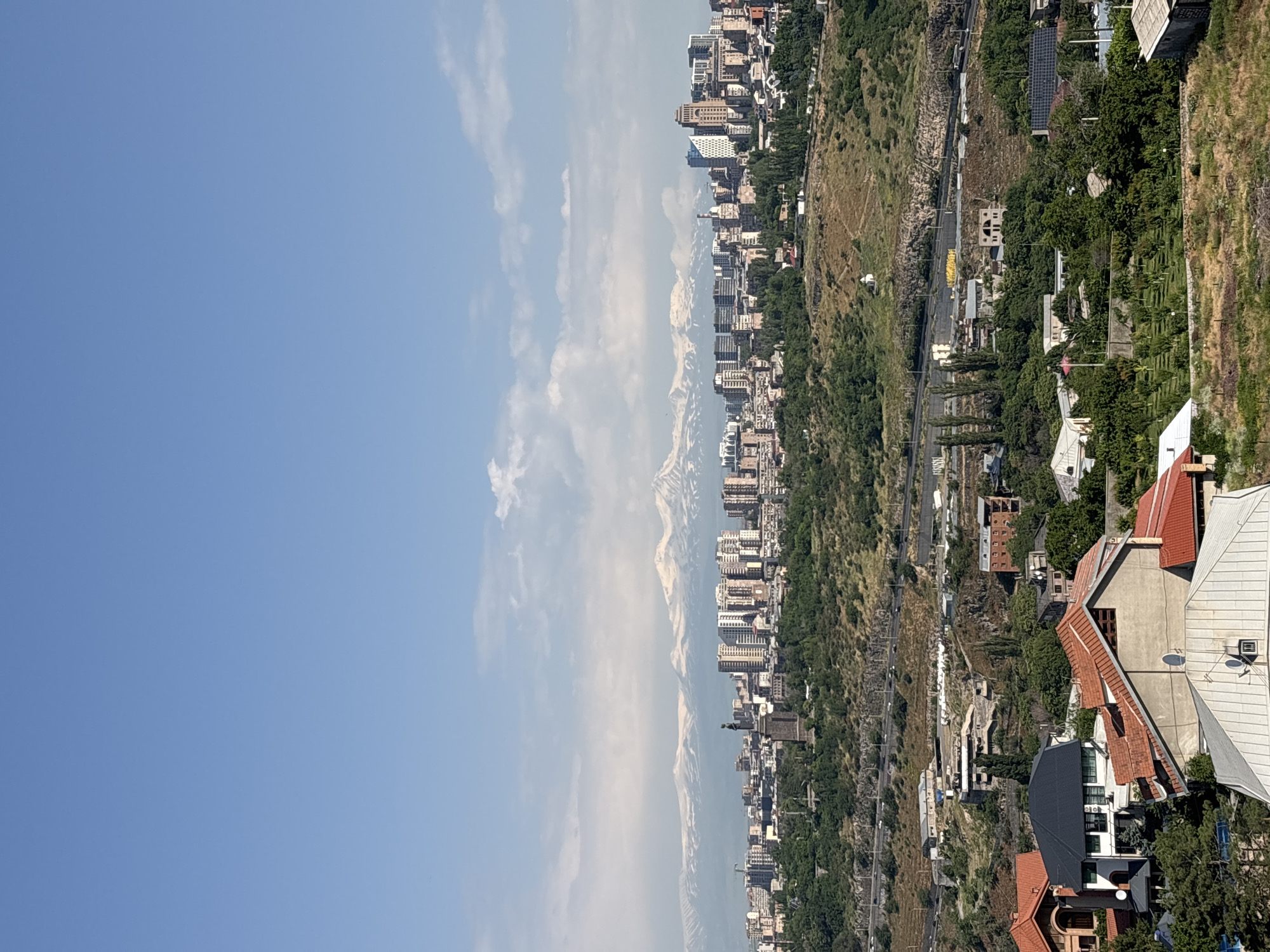This is a wide-angle, eye-level, outdoor, daytime photograph taken from a high vantage point overlooking a city. The sky is a clear, pale blue with some thin, wispy clouds visible near the horizon. In the distance, a majestic mountain range with snow-capped peaks dominates the background, suggesting a cool climate or high altitude.
In the mid-ground, the city of Yerevan, Armenia, unfolds. A dense cluster of modern buildings, ranging from mid-rise apartment complexes to taller skyscrapers, forms the urban core. These structures exhibit a variety of architectural styles and materials, with predominantly light-colored facades and numerous windows. A prominent statue, likely a monument, stands amidst the city buildings. Below the city, a verdant, tree-covered hillside slopes downwards. A road with some traffic is visible winding along the hillside.
The foreground of the image features a residential area with houses. These homes are characterized by red-tiled roofs, and some have visible solar panels on their roofs. The terrain in the foreground is a mix of dry grass and some scattered trees. The overall impression is a panoramic view of a city with a striking natural backdrop.