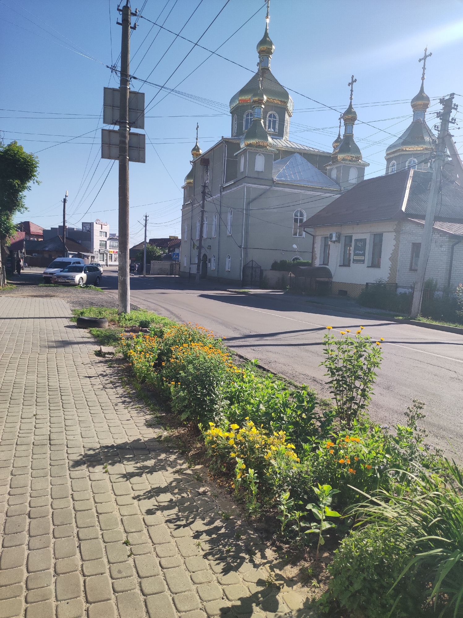 A low-angle, eye-level shot captures a sunlit street scene in an urban environment. The foreground is dominated by a cobblestone sidewalk on the left, casting long shadows from an unseen tree. Adjacent to the sidewalk is a narrow strip of green grass and flowerbeds filled with yellow and orange flowers, with a discarded tire to the left of the flowers. The street itself is paved and curves slightly to the right.
In the middle ground, a multi-domed, grey-colored church with golden cupolas stands prominently on the right side of the street. A smaller, older brick building with a red roof is situated in front of the church. Across the street, on the left, a white van and a silver car are parked, with a row of buildings behind them, including a modern, multi-story building with signs. A utility pole with numerous wires and two square signs is positioned on the sidewalk.
The background shows a clear, bright blue sky with some overhead wires. The scene appears to be a quiet, sunny day in a town or city, likely in a region with Eastern Orthodox architecture. There are no discernible people or animals in the immediate foreground or middle ground.
