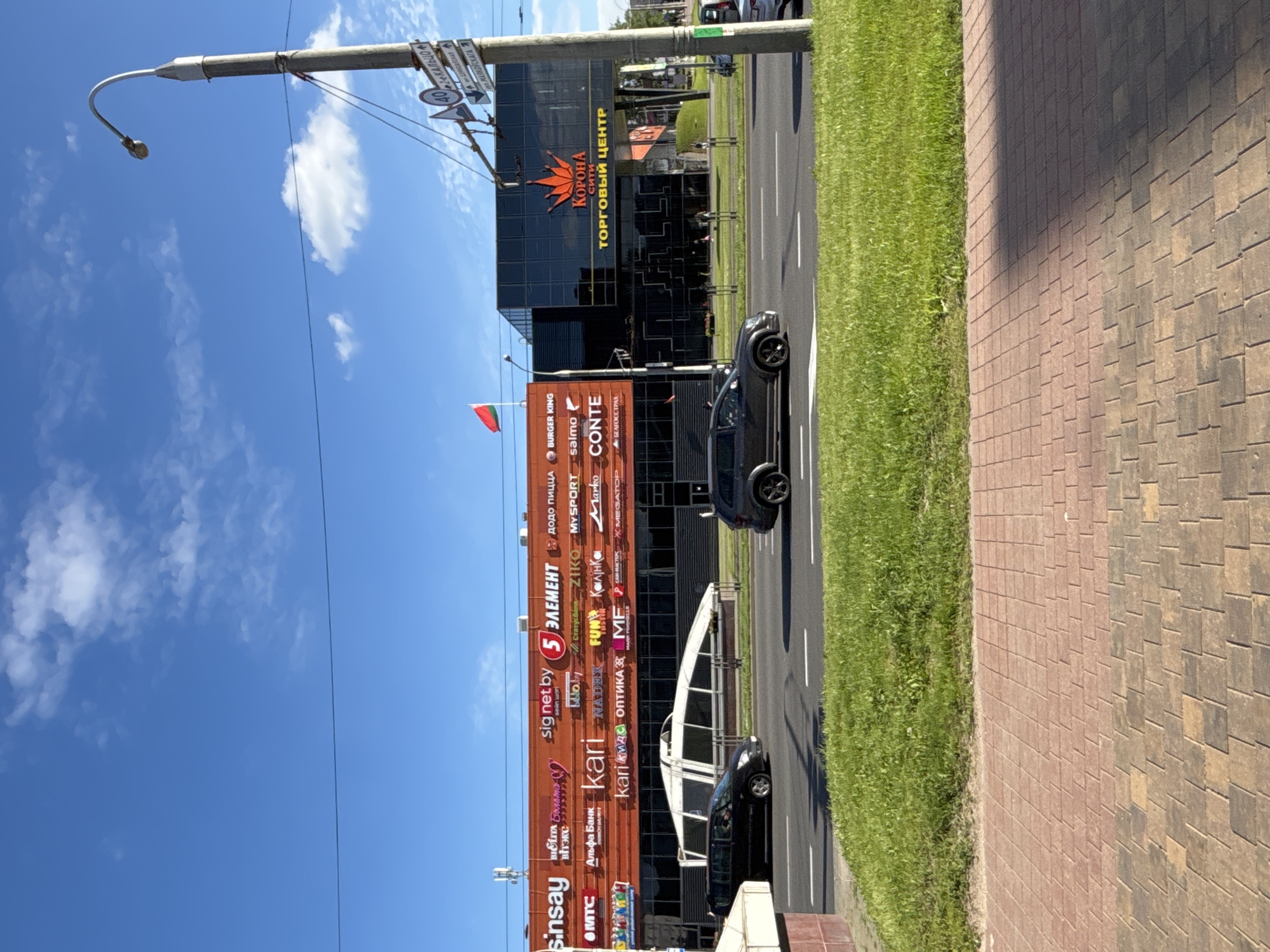 The image depicts a street scene in Minsk, Belarus, on a clear day with scattered clouds. A large shopping center dominates the background, characterized by an orange-colored facade on the left section adorned with various brand logos, including "Sinsay," "5 ЭЛЕМЕНТ," "MTC," "Alpha Bank," and "Kari." The right section of the shopping center features a modern black glass facade with a prominent red crown logo and the text "КОРОНА СИТИ ТОРГОВЫЙ ЦЕНТР" (Korona City Shopping Center).
In the middle ground, a multi-lane road is visible with several cars. A black minivan is driving in the left lane, and a dark gray SUV is in the right lane. A grassy median separates the road from a sidewalk made of red and brown paving stones. A lamppost stands on the right side of the image, equipped with streetlights and traffic signs, one of which indicates a speed limit of 40. A Belarusian flag flies from a pole on top of the shopping center. The overall environment suggests a commercial area within the city.