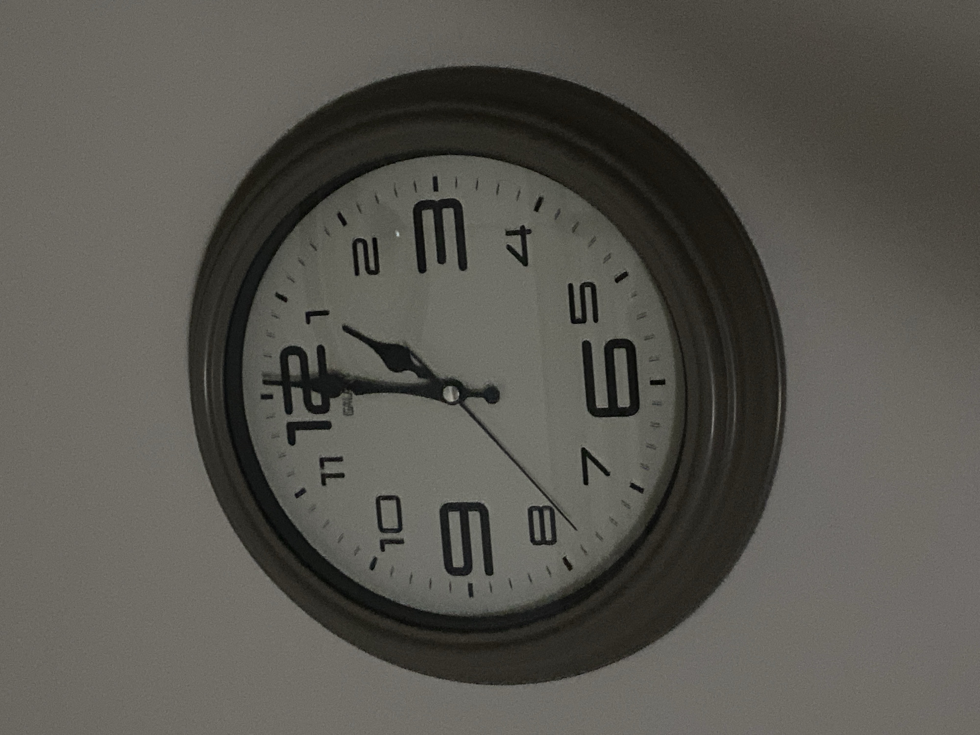 A wall clock is displayed against a neutral-colored wall. The clock has a round, dark frame and a white face with black Arabic numerals. The hour hand is pointing slightly past the 12, and the minute hand is pointing at the 3. There is a small logo that says "GAUDI" in the center of the clock face. The image appears to be taken in the city of Kocasinan, Turkey, but no specific location within Kocasinan is identifiable from the image. The scene is static and does not contain any people or discernible actions. The lighting is dim, suggesting it may be indoors or during low-light conditions.