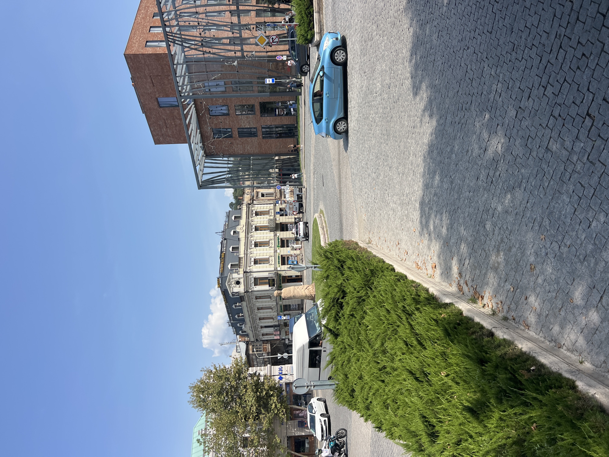 This is a street-level, eye-level, full shot of a paved intersection in Tbilisi, Georgia, on a clear day with a blue sky and some scattered clouds. In the foreground, a well-maintained, lush green hedge forms a circular median. The cobblestone street is visible throughout the scene, with cars driving and parked.
On the left side of the frame, a white van is stopped at the intersection, partially obscured by the hedge. A white sedan is driving away from the viewer, and a motorcyclist is riding towards the viewer. Behind them, several shops and buildings line the street, one with a green roof.
In the center of the intersection, a statue of a person stands on a pedestal. Further back, an older, ornate building with a multi-story facade and a prominent roofline is visible. A sign on this building reads "MONUMENT 1888".
On the right side of the frame, a modern, multi-story brick building with a distinctive metal-framed glass facade is prominent. Cars are driving in front of this building, including a dark gray SUV and a bright blue hatchback. Several pedestrians are visible walking on the sidewalk in front of the modern building.
The overall scene depicts a busy urban intersection with a mix of old and new architecture.