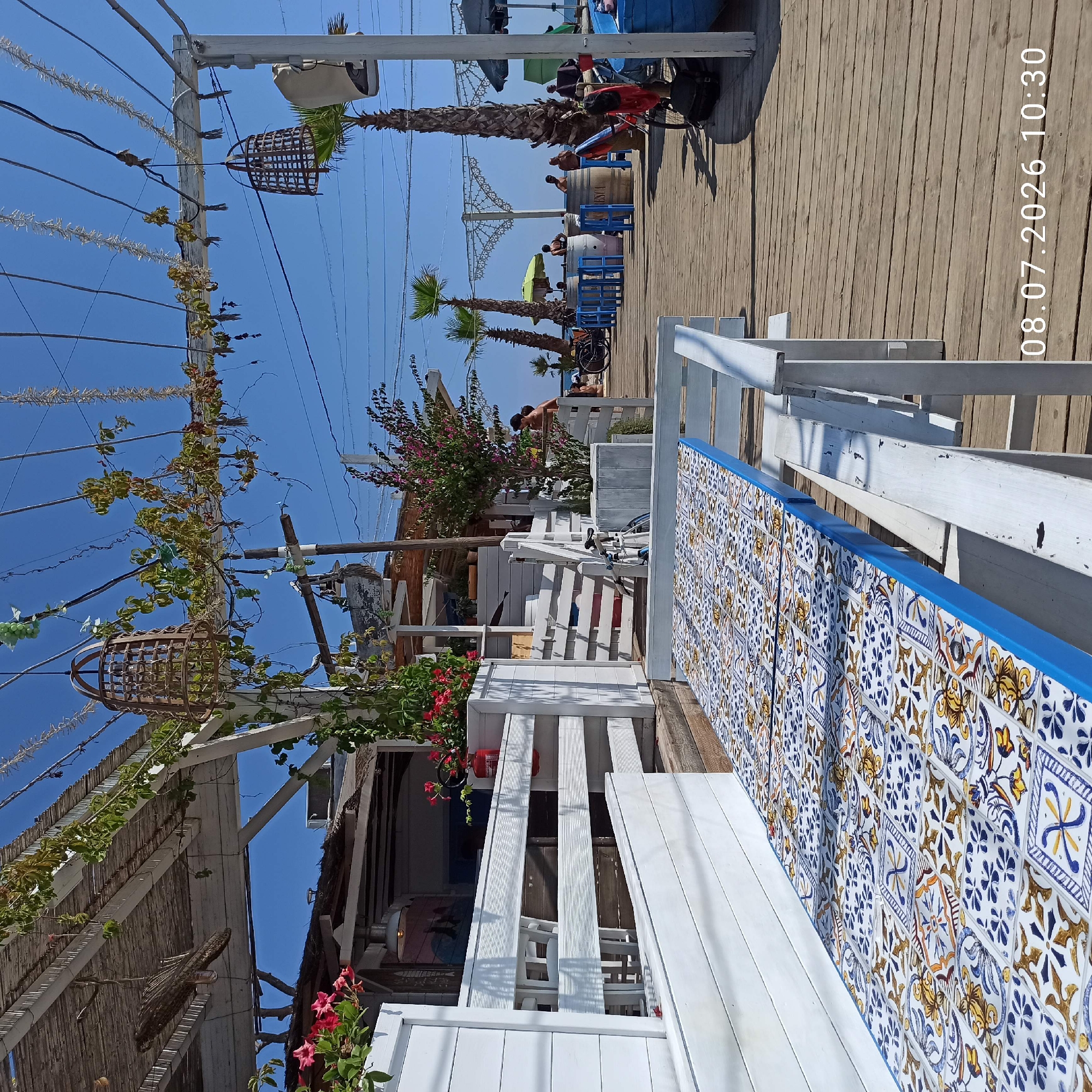 This image depicts a sunny outdoor seating area, likely a cafe or restaurant, situated on a wooden boardwalk by the sea. In the foreground, a white bench with a table covered in a decorative tiled pattern is visible. The tiles feature a blue, yellow, and white design. Behind the bench, a white wooden fence encloses more seating areas. Further back, there is a structure with a thatched roof and white wooden supports, adorned with hanging baskets and string lights. Several palm trees are scattered throughout the scene. In the background, people are seen relaxing on the beach and near the seating area, some standing and some sitting. A bicycle is also visible. The sky is clear and blue. The image is dated 08.07.2026 10:30. Based on the architectural style, the presence of palm trees, and the general ambiance, this scene is likely located in Trani, Italy, possibly along its waterfront promenade or a beachside establishment.