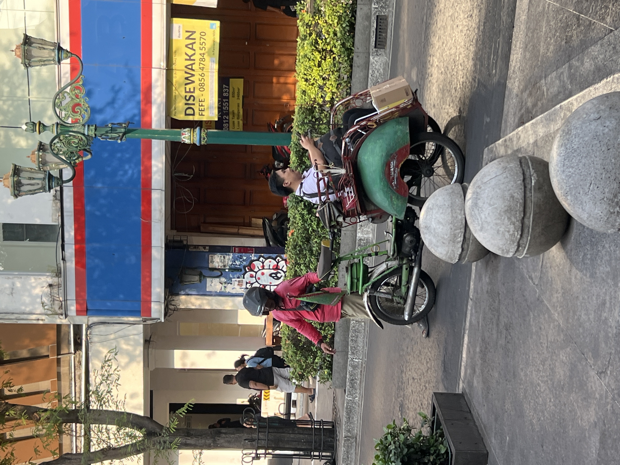 In a street scene in Yogyakarta, Indonesia, a pedicab driver, wearing a pink long-sleeved shirt, tan pants, and a black helmet, stands next to his green and red pedicab. The pedicab has a passenger, a man in a white and black collared shirt, seated in the back and looking at his phone. A brown cardboard box is secured to the back of the pedicab. In the background, a building with a blue and red horizontal stripe design is visible, along with a green street lamp with ornate details. Several people walk on the sidewalk in the distance. The street is paved, and a row of decorative spherical planters lines the sidewalk in the foreground. The overall lighting suggests it is daytime.