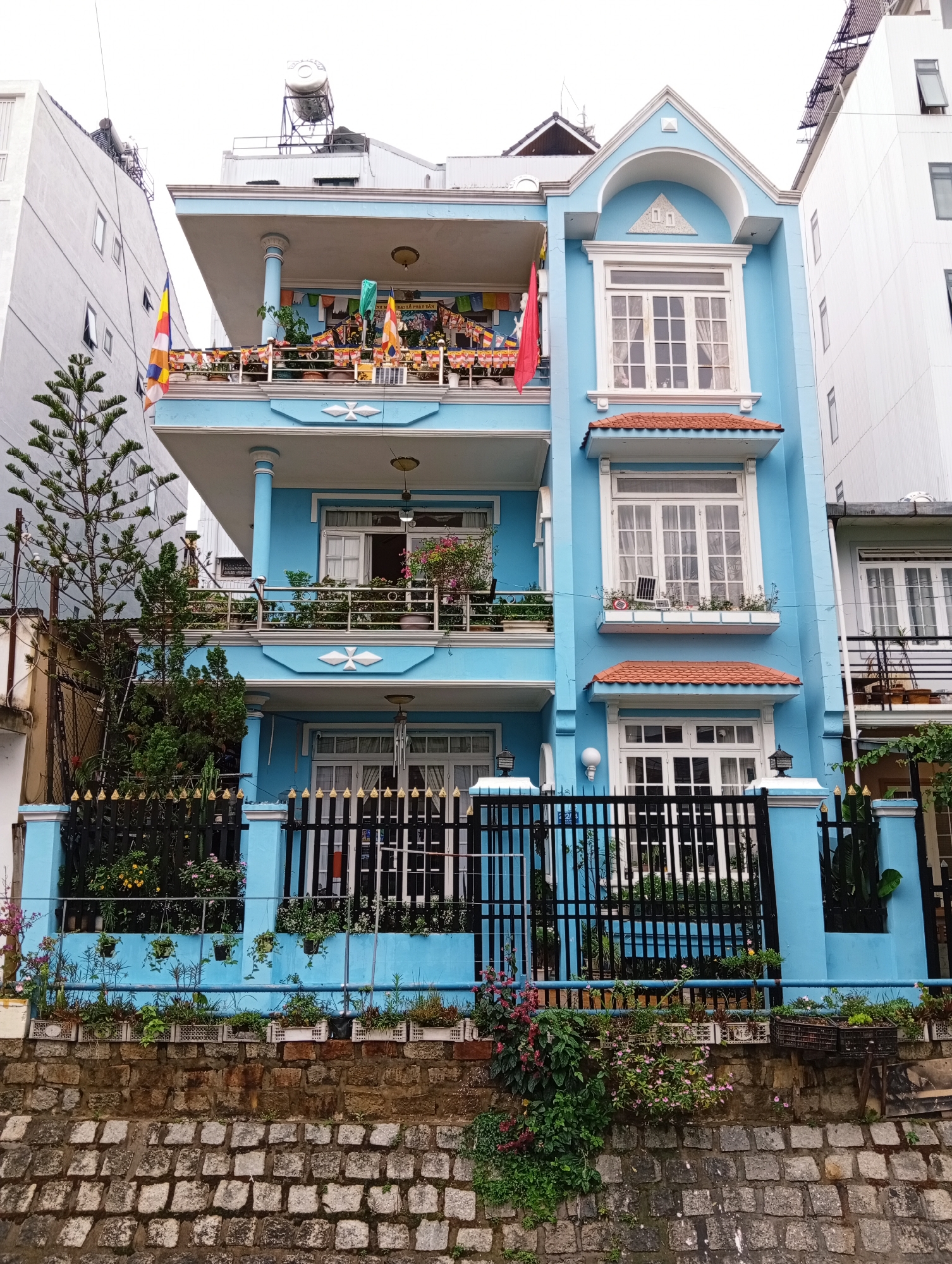 The image shows a three-story blue building with white trim, located in Da Lat, Vietnam. The building has multiple balconies with railings, decorated with various plants and flags. The front of the building features large windows with white frames and red-tiled awnings. A black metal fence surrounds the building at street level. The building is situated between two taller, nondescript buildings. A stone wall borders the bottom of the building, with more plants and flowers growing along it. The sky is overcast.