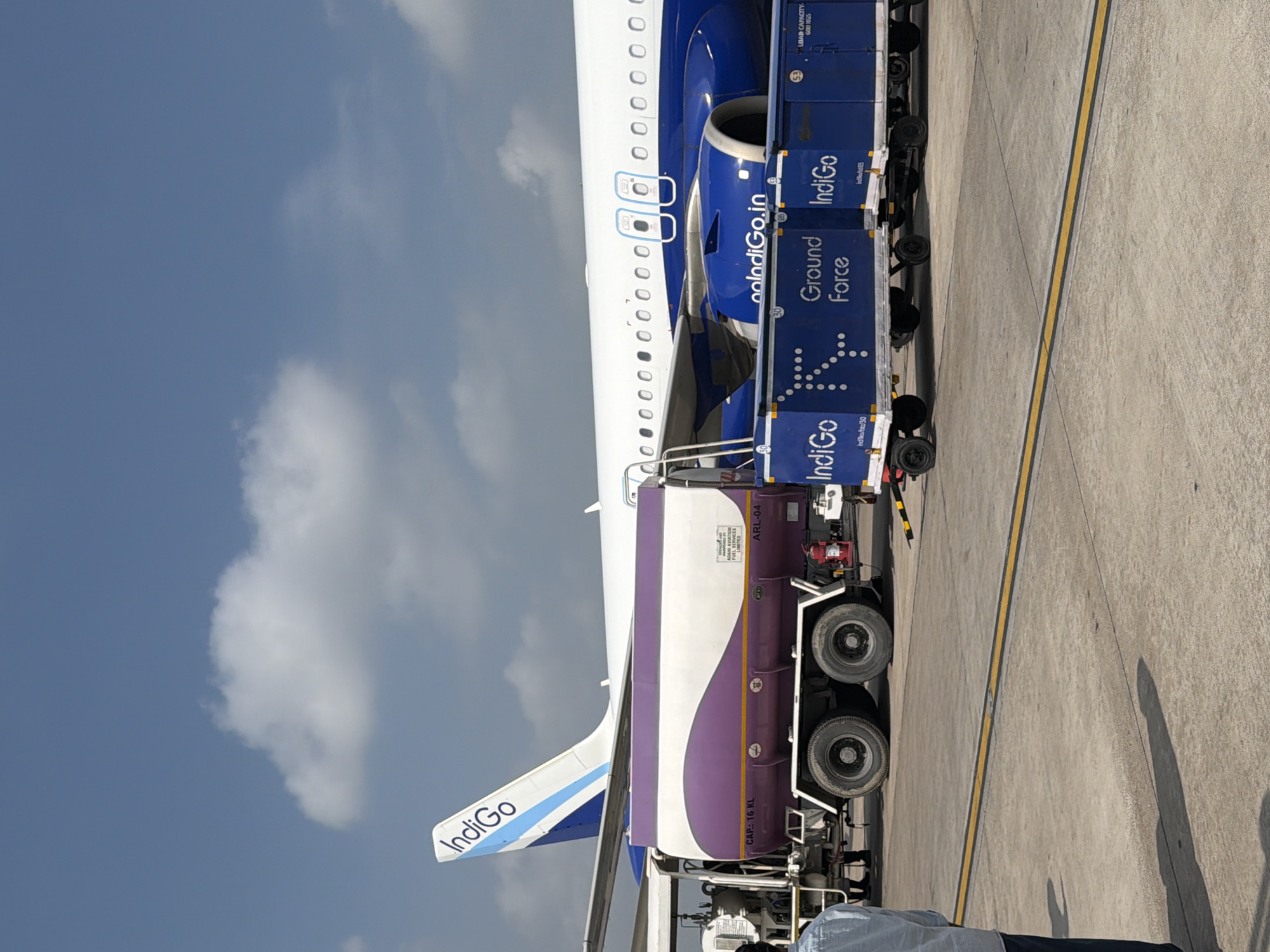 An Indigo Airlines airplane is parked on a tarmac. A fuel truck with a purple and white tank is positioned next to the plane. Several blue baggage carts with "Indigo Ground Force" written on them are also present. A person in a light blue shirt and dark pants stands to the left of the frame. The sky is mostly clear with some scattered clouds. The scene appears to be at an airport in Vikram Nagar, India.