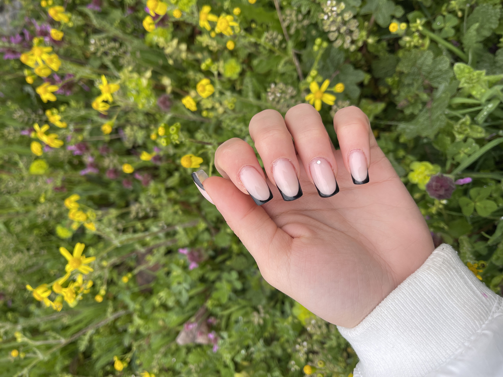 A hand with a fresh black-tipped French manicure is prominently displayed against a blurred background of lush green vegetation dotted with small yellow and purple flowers. The hand is light-skinned, and the nails are well-manicured. A small, singular bead adorns one nail. The person is wearing a white long-sleeved shirt or jacket, with only the cuff visible at the bottom right. The background suggests a natural setting, possibly a garden or meadow. The location within the city of Veliko Tarnovo, Bulgaria, is not discernible from the visual information provided.