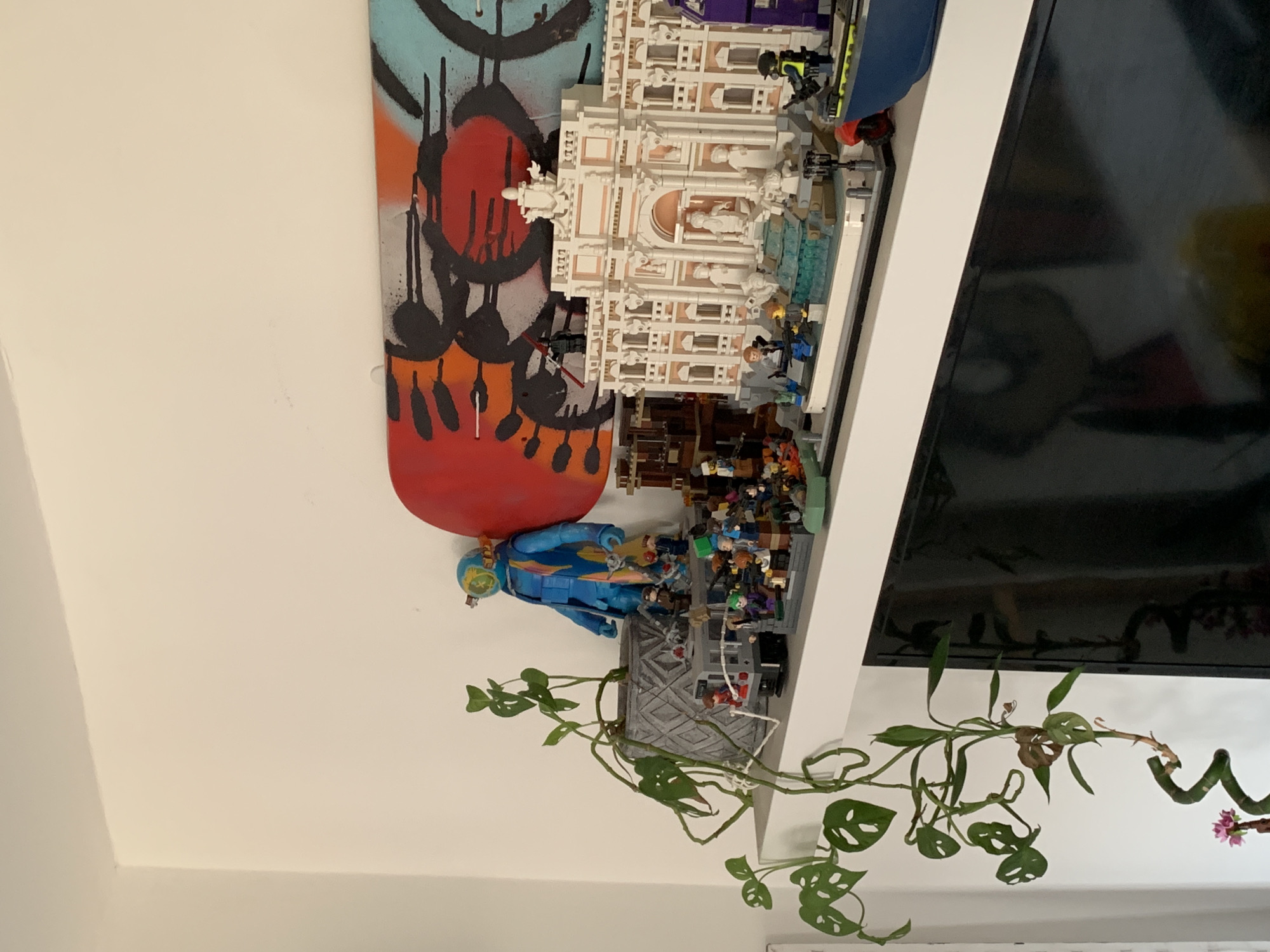 This image captures an indoor display, rotated 90 degrees counter-clockwise, suggesting items arranged on a shelf against a white wall. On the left, a skateboard or longboard stands upright, featuring vibrant graffiti art with red, orange, black, and light blue elements. Adjacent to it, a large, intricate LEGO architectural model, possibly depicting a historical building like the Trevi Fountain, dominates the center. The model is primarily white or cream-colored, showcasing elaborate details and several small LEGO minifigures scattered around and within it.
To the left of the LEGO model, a prominent blue and teal figure, resembling an astronaut or diver in a specialized suit, stands. Further to the right, a large, black flat-screen television is visible, with its reflective surface showing faint, indistinct reflections of the room. Hanging and trailing from the display surface is a lush green plant, likely a Monstera Adansonii (Swiss Cheese Plant), characterized by its leaves with distinctive natural holes. Some leaves appear to have brown, dried areas. On the far right, another plant features a uniquely spiraled stem, possibly a lucky bamboo, with a few pink artificial flowers attached. The scene is brightly lit, with no specific time of day discernible. No activity or human presence is directly depicted, and no visible text is present.
