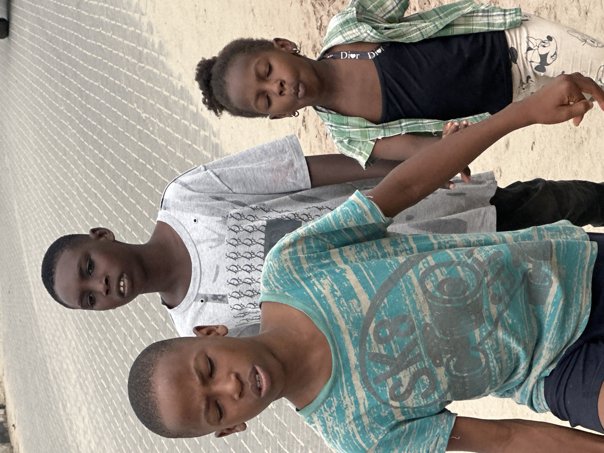 In Badagry, Nigeria, three children walk along a paved surface. In the foreground, a young boy wearing a turquoise and white striped t-shirt and navy shorts walks towards the camera, looking down. Behind him, a slightly older boy in a grey t-shirt with a repeating pattern walks, looking forward. To the right, a young girl in a green plaid shirt over a black tank top and patterned leggings walks, closing her eyes and pursing her lips. The ground is a light-colored pavement with diagonal lines.