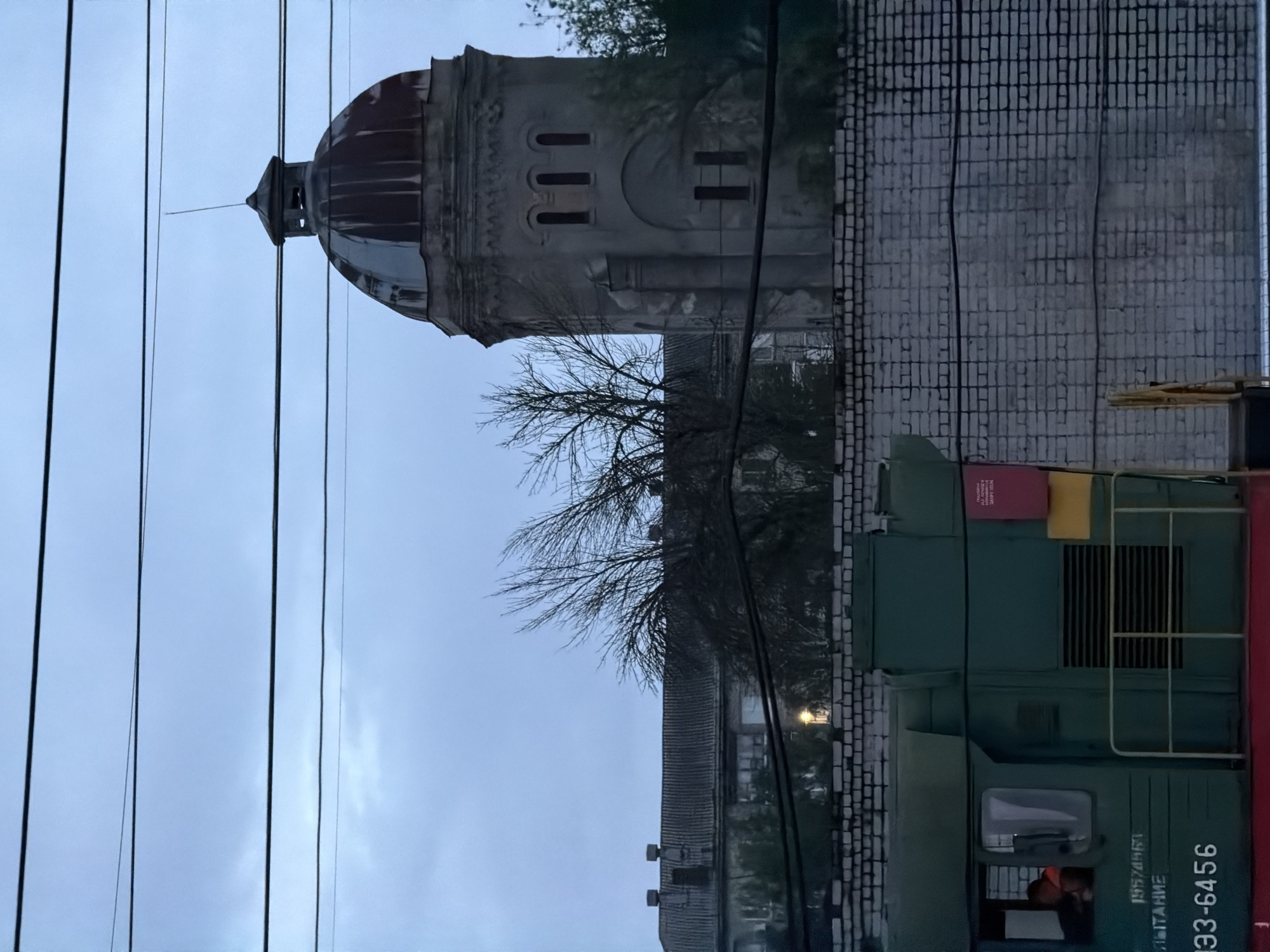 The image shows a water tower, a brick wall, and a green and red train locomotive in Rtishchevo, Russia. The tower has a dome-shaped top and several arched windows. Bare tree branches are visible in front of the tower and brick wall. Power lines run across the sky. The train locomotive has the number "933-6456" printed on its side. A person is visible through the train cab window. The overall lighting suggests it is dusk or dawn.