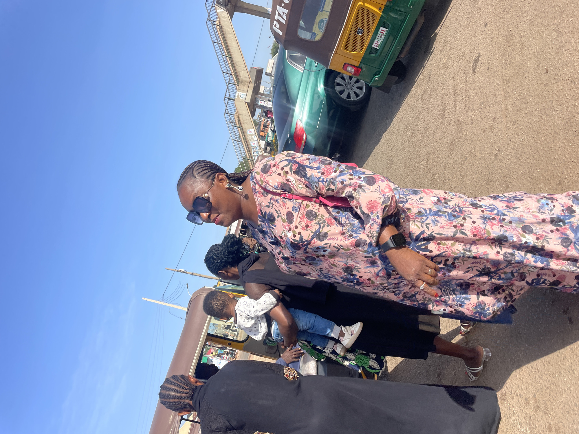 The tilted image captures a vibrant, sunny outdoor scene in Gura Top, Nigeria, depicting several individuals on what appears to be a street or pathway. In the foreground and central to the image is a woman walking, dressed in a long-sleeved floral dress featuring pink, blue, and black patterns. She wears sunglasses, has intricately braided hair, a smartwatch on her left wrist, and a small pink cross-body bag. Her feet, adorned with clear-strapped sandals with white detailing, are visible on the paved ground.
Behind her, another woman in a black dress is seen carrying a young child, who is dressed in blue jeans and a white long-sleeved top. Part of another person in black clothing is visible in the bottom foreground of the image.
The background reveals a bustling urban environment under a clear blue sky. A green and yellow tricycle, often called a "keke napep," is prominently featured, with the license plate "DAG-786JW" clearly visible on its rear. Partially visible text on its side reads "PTA-". A green sedan is located behind the tricycle. In the upper background, a concrete pedestrian bridge or overpass spans above the road, and utility poles with wires are also visible. The strong sunlight casts distinct shadows, indicating a bright, clear day, likely during the mid-morning or afternoon. The overall scene suggests routine activity and movement in a lively, sunny setting.