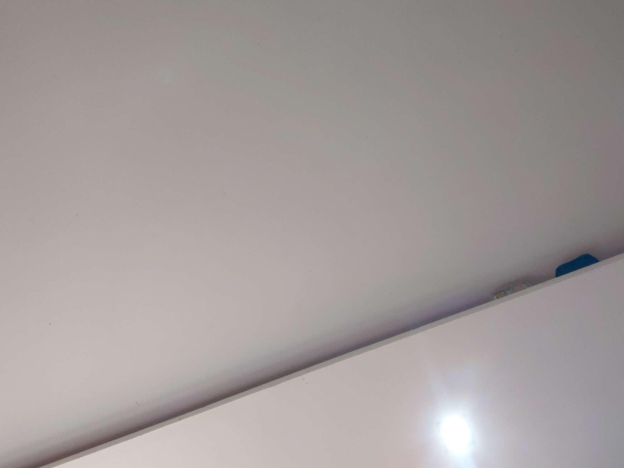 The image provides an upward-angled view within an interior space, predominantly featuring two large, light-colored surfaces. The upper portion shows a smooth, uniform ceiling or upper wall, which is off-white or light grey. Below this, separated by a narrow linear gap, is another flat, light-colored surface, likely the top of a wall or a tall piece of furniture such as a cabinet or wardrobe.
In the gap between these two surfaces, towards the far right, a small, distinct blue object is partially visible. To its left within the same gap, there is some lighter, possibly yellowish or white material, which could be insulation or accumulated debris.
A very bright, circular light source or reflection creates a prominent glare in the lower right section of the image, indicating the presence of an active artificial light fixture, although the fixture itself is not fully depicted. The surfaces appear clean and well-maintained.
There are no people or any discernible activity taking place in the scene, which captures a static architectural detail. The time of day cannot be determined, as the illumination primarily comes from the artificial light source. No text or specific decorative elements are visible. The image offers general interior details without specific visual cues to identify its location as Santa Ana, Venezuela, beyond the provided context.