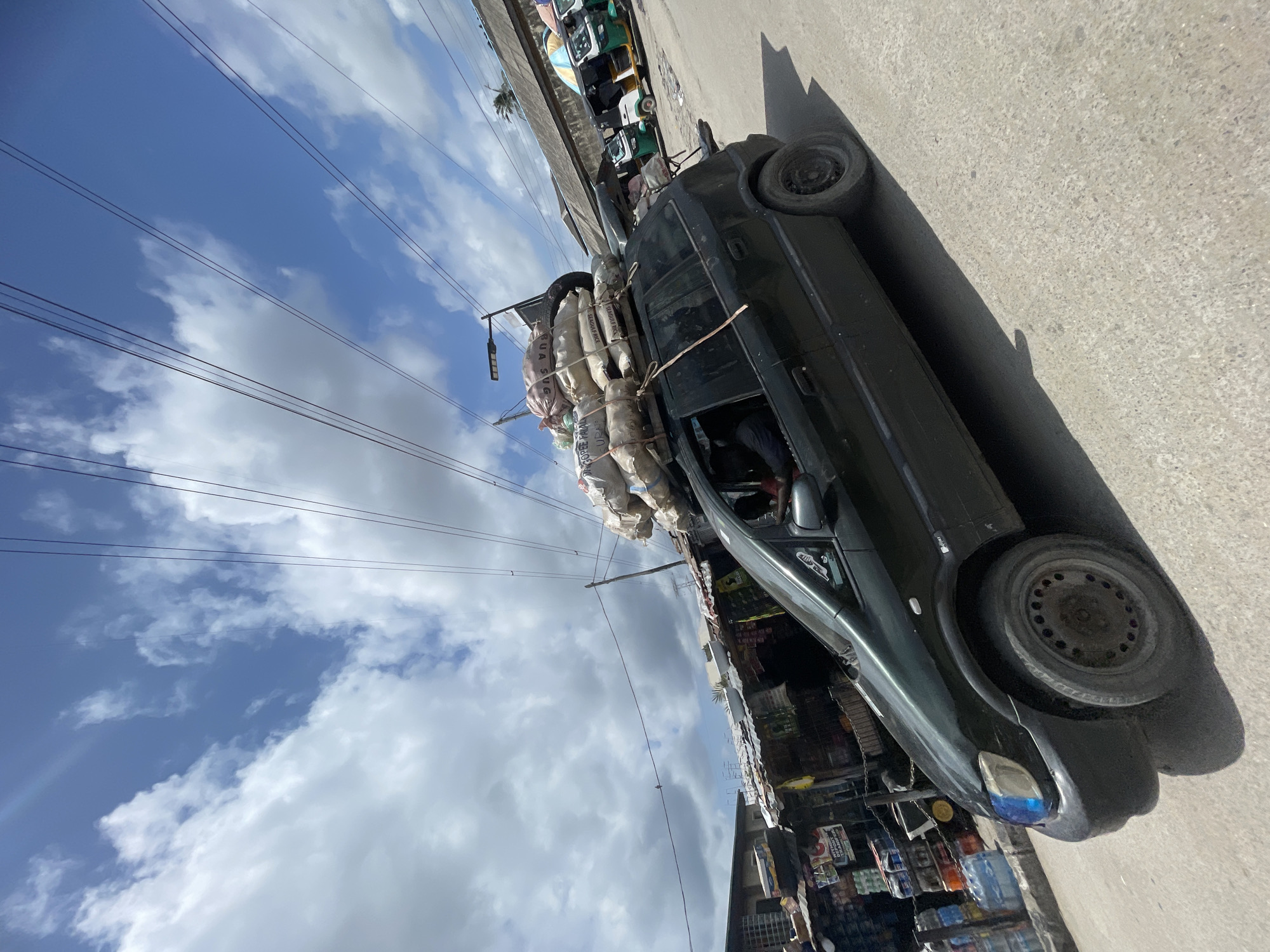 A heavily laden black car is parked on a street in Ado, Nigeria. The car's roof is piled high with bags secured with rope. An overhead power line is visible against a backdrop of blue sky with scattered white clouds. In the background, there are buildings and motorized rickshaws. Goods are displayed outside of stores next to the car.